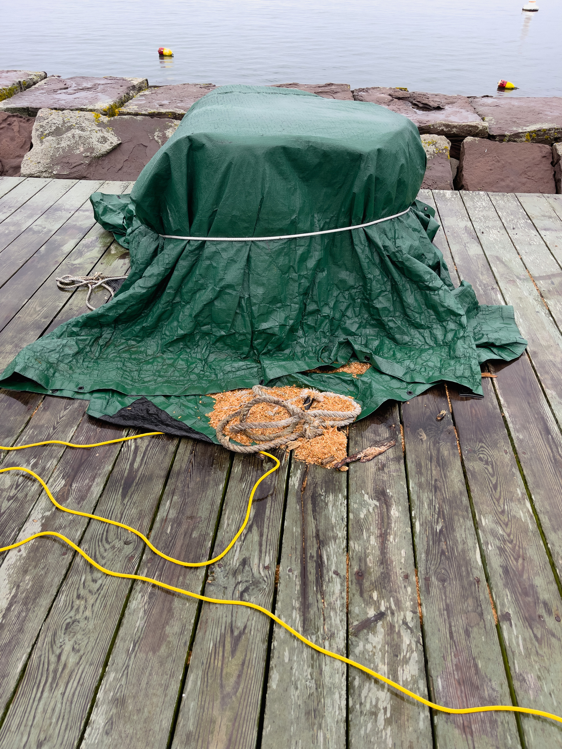 Green tarp over rectangular objects with rope and yellow power chord on dock boards in front.