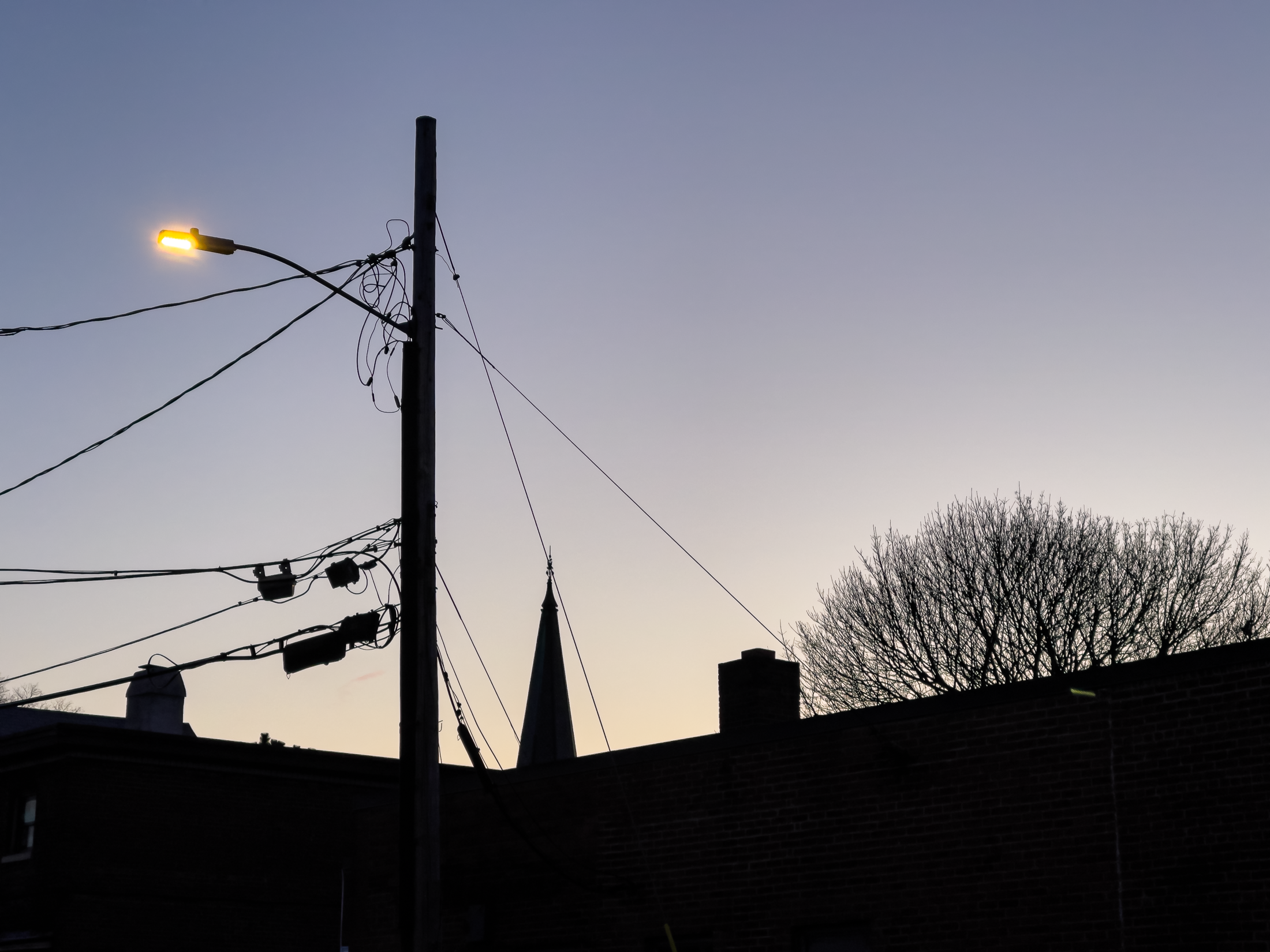 Building, streetlight, utility pole, wires and church steeple silhouetted against the morning sky.