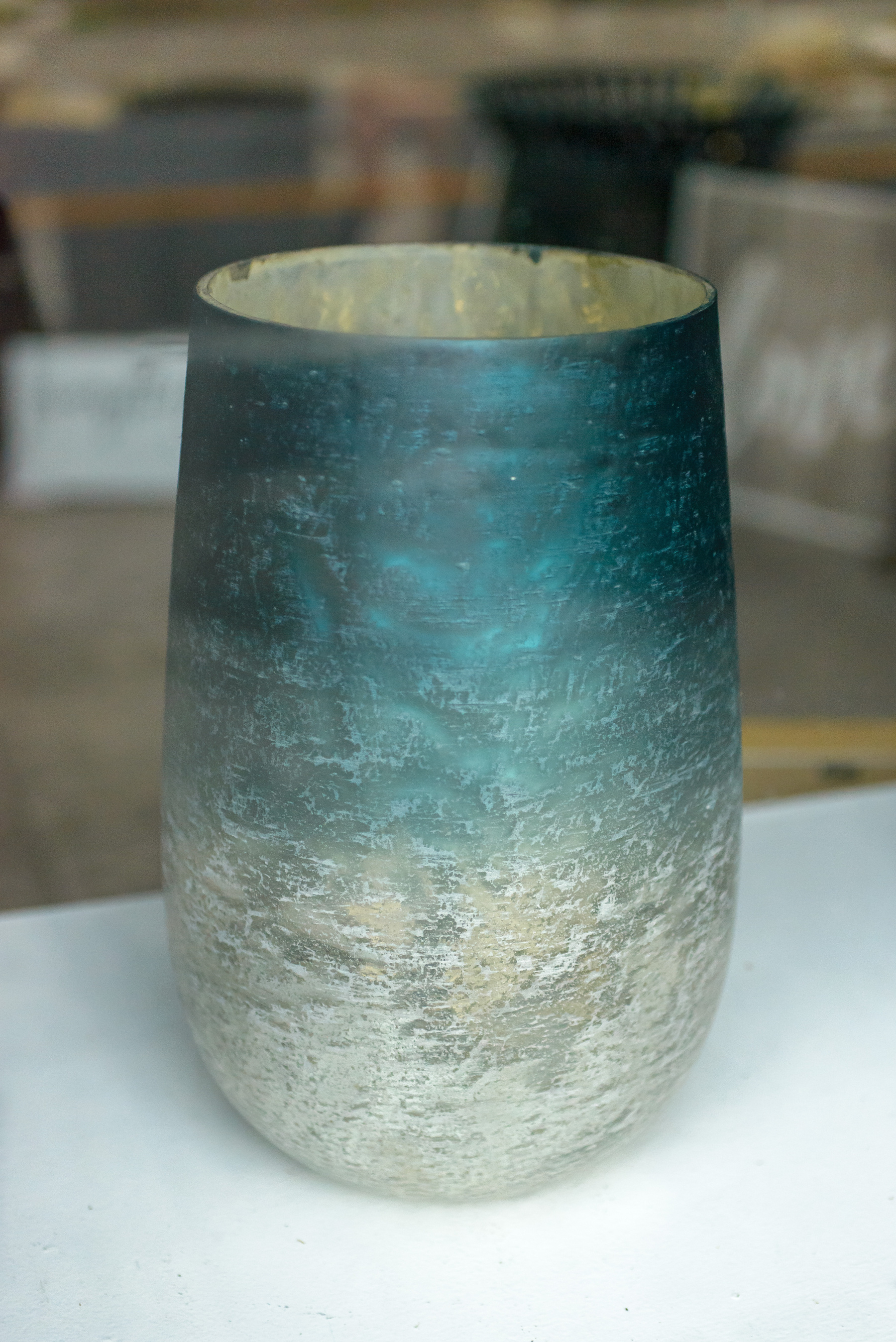 Glass antique vase in a shop window with reflective silver inside and a gradation of dark to light turquoise green from top to bottom. Vase is circular with sides widening out uniformly two thirds of the way down, then gently curving down and in to the narrower base. 