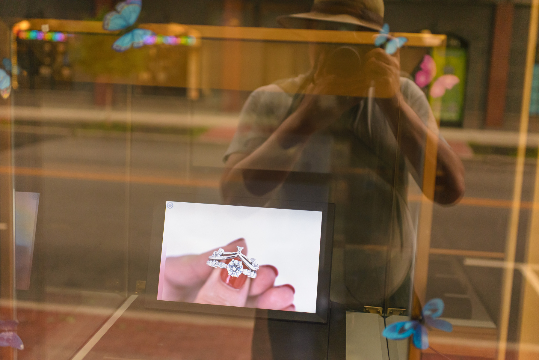 Shop window with display screen showing a woman hand with red nails holding a diamond ring. Photographer is reflected in the window along wit a streetscape.