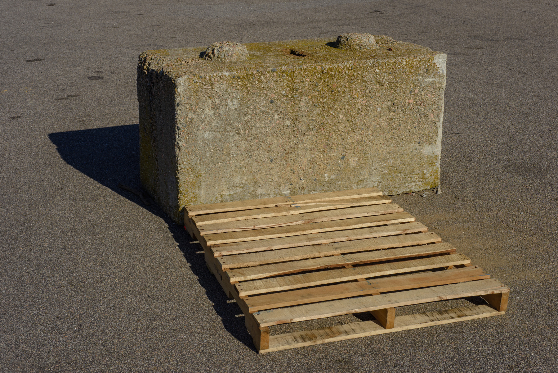 Concrete barrier with wooden pallet on the ground in front of it, on an asphalt paving surface.