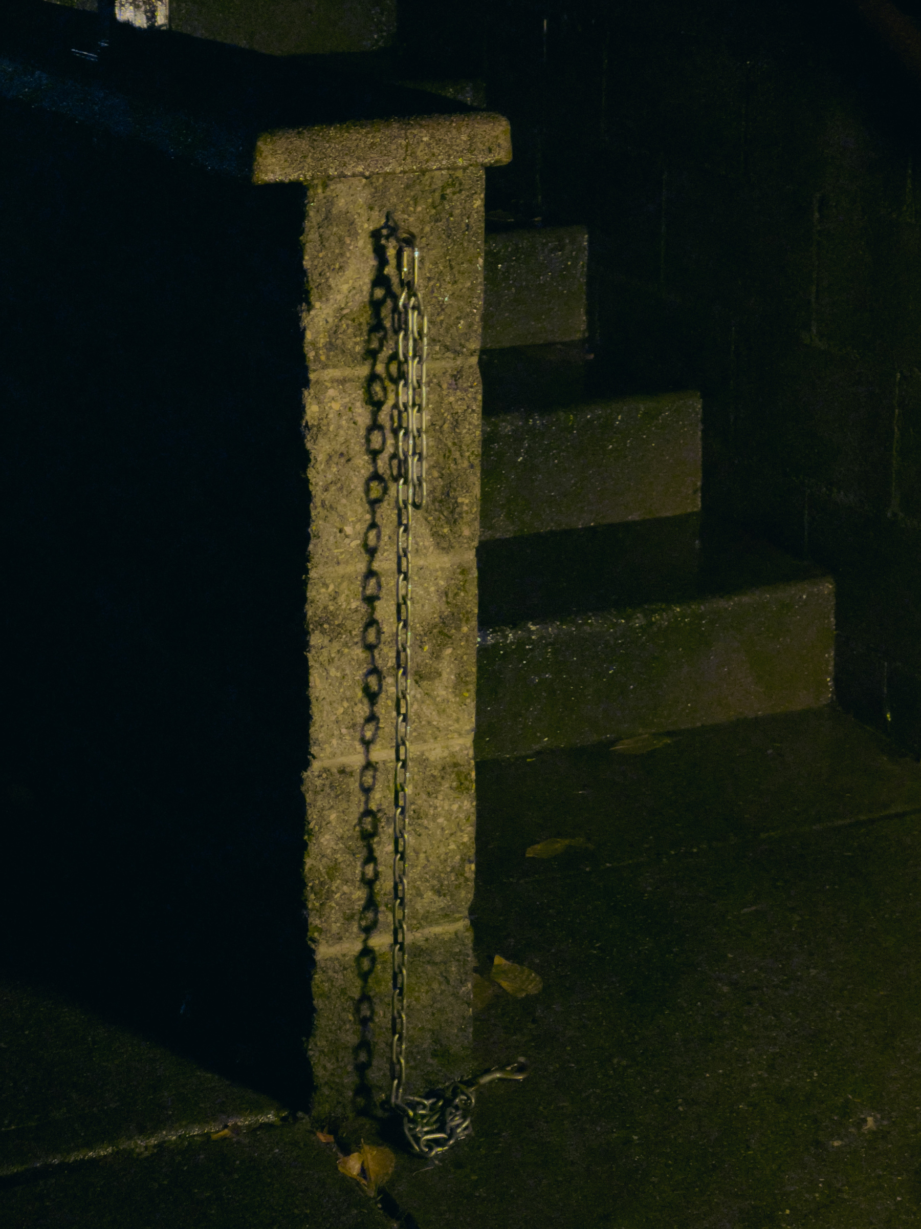 End of low height wall with chain dangling from top to pavement below, concrete stairs in shadow beyond, scene illuminated by unseen security light to the right 