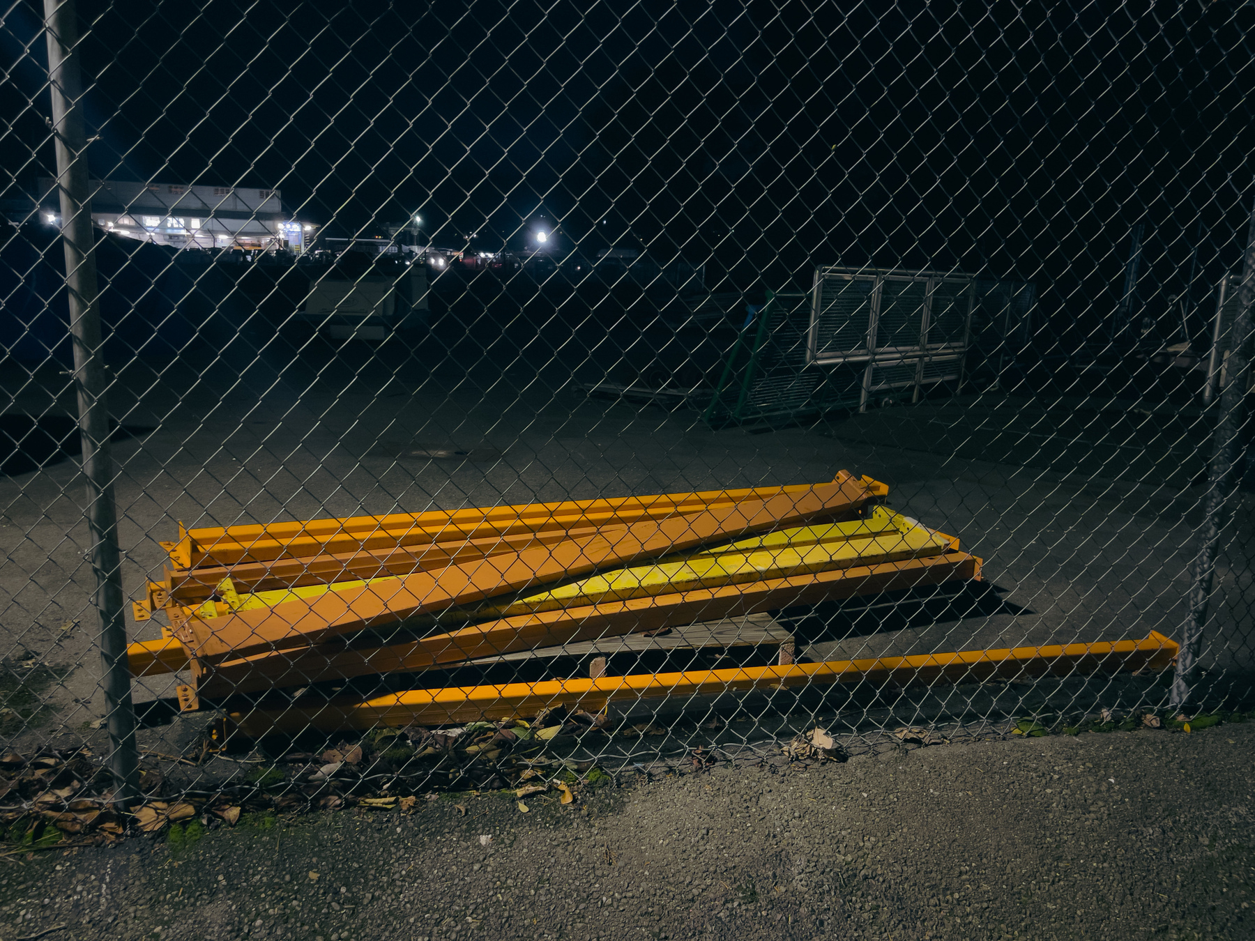Rusted steel framing components on ground behind chain link fence, scene illuminated by security lights.