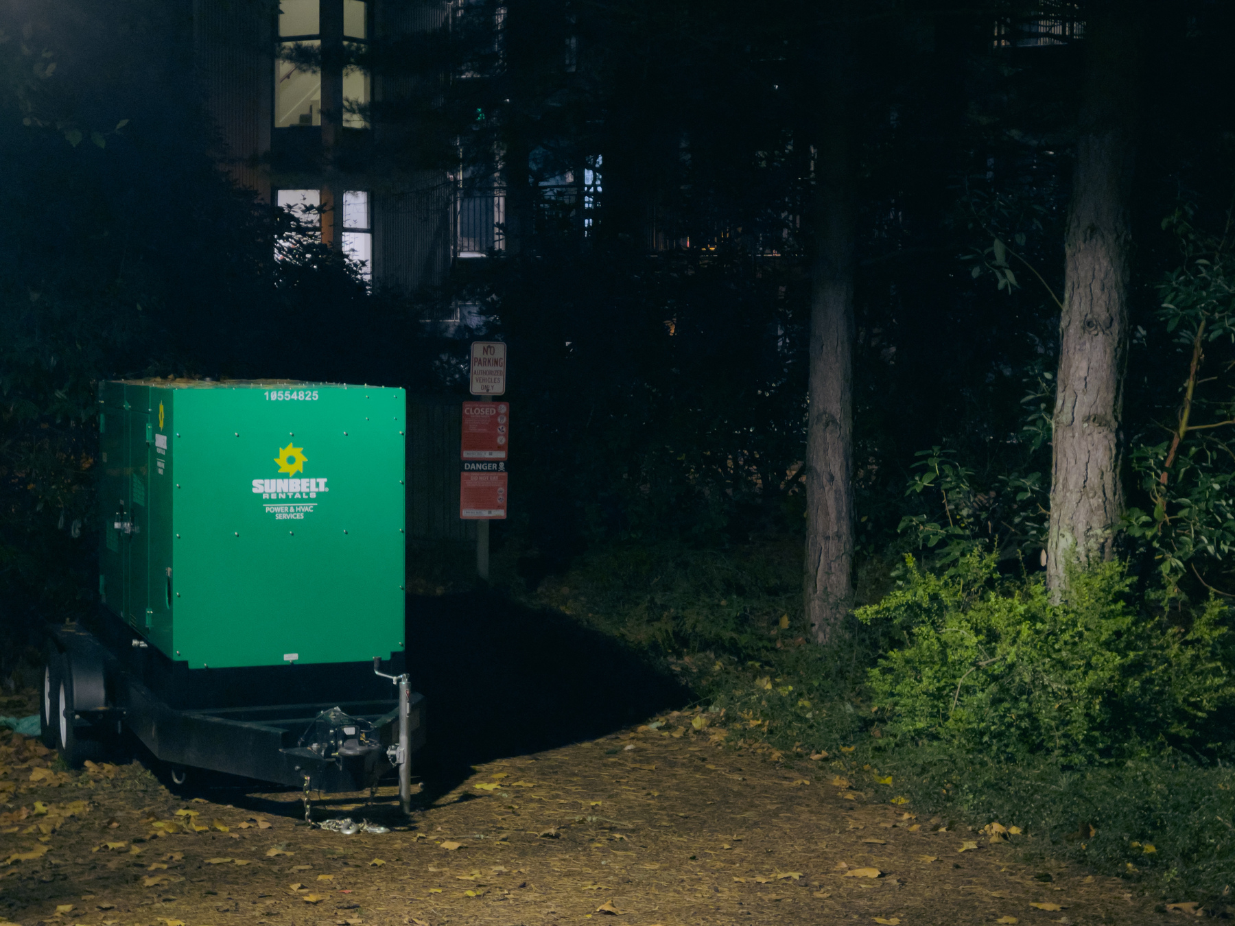 Portable generator in clearing between trees, illuminated windows of multi story building in distance, scene illuminated by security lights.