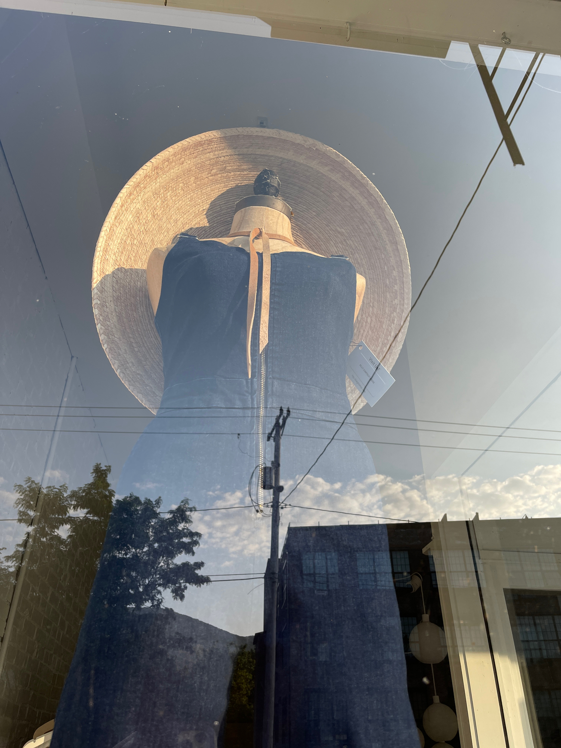 Women’s bluejeans jump suit with large wide brim woven hat on mannequin in a shop window. Sky clouds and buildings reflection overlaid.