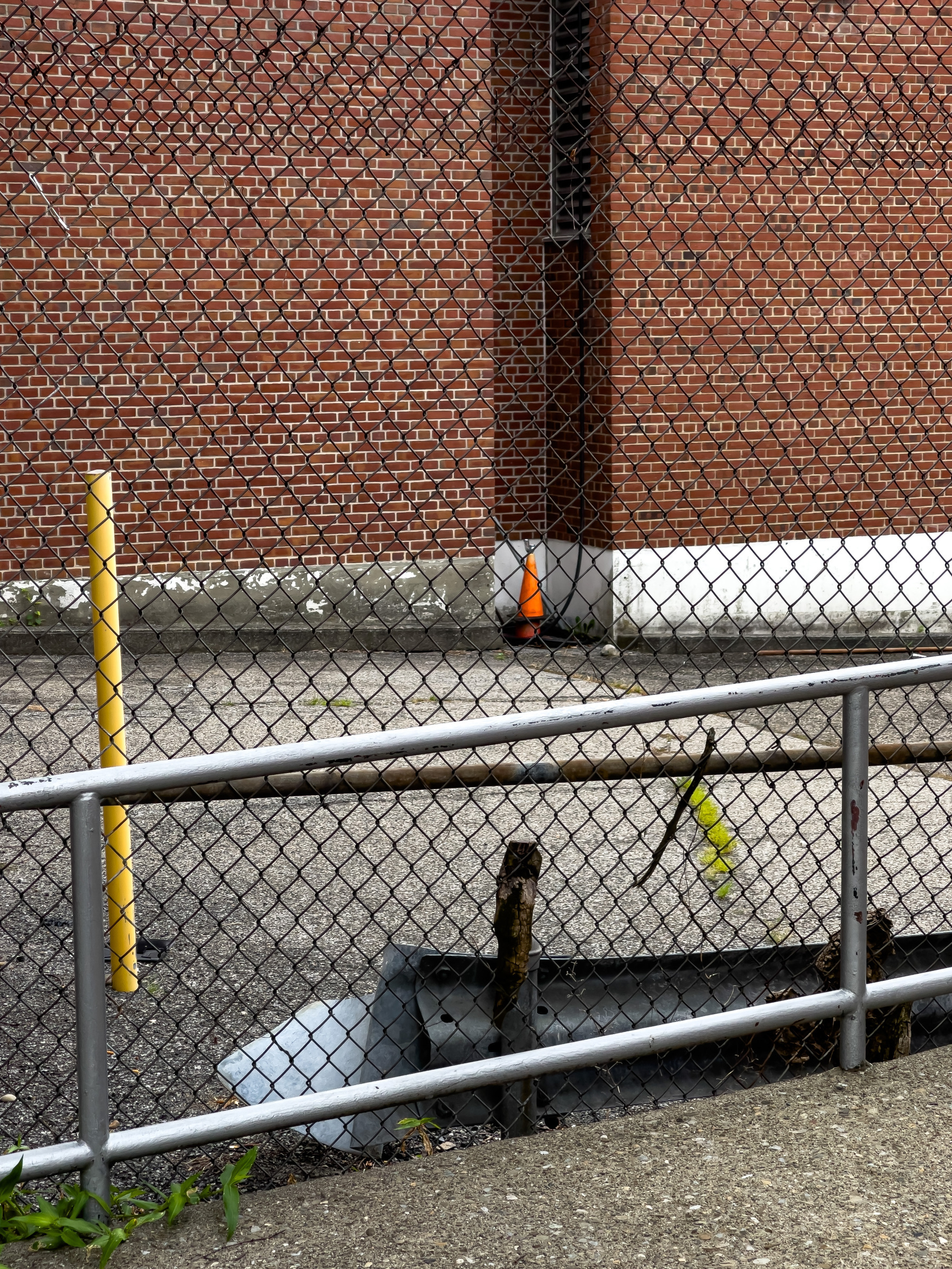 Streetscape, side of brick building in the background, metal railing and chain link fence in foreground, vertical yellow pipe vehicle barrier in the near mid ground on the left.