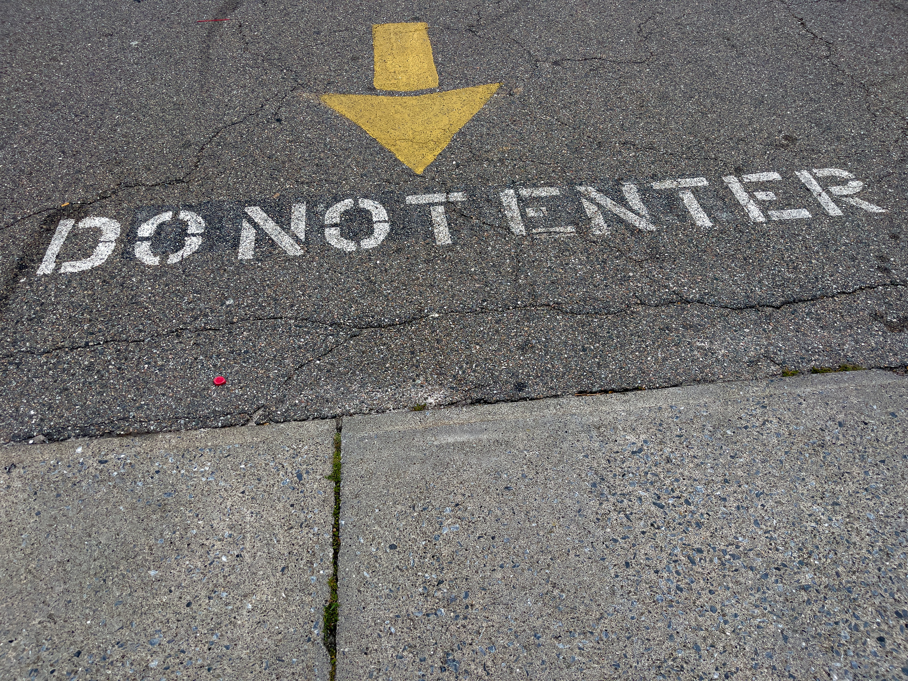 Yellow arrow painted on asphalt parking lot in middle top of frame pointing towards the bottom of the frame with the words “DO NOT ENTER” painted in white below the tip of the arrow.