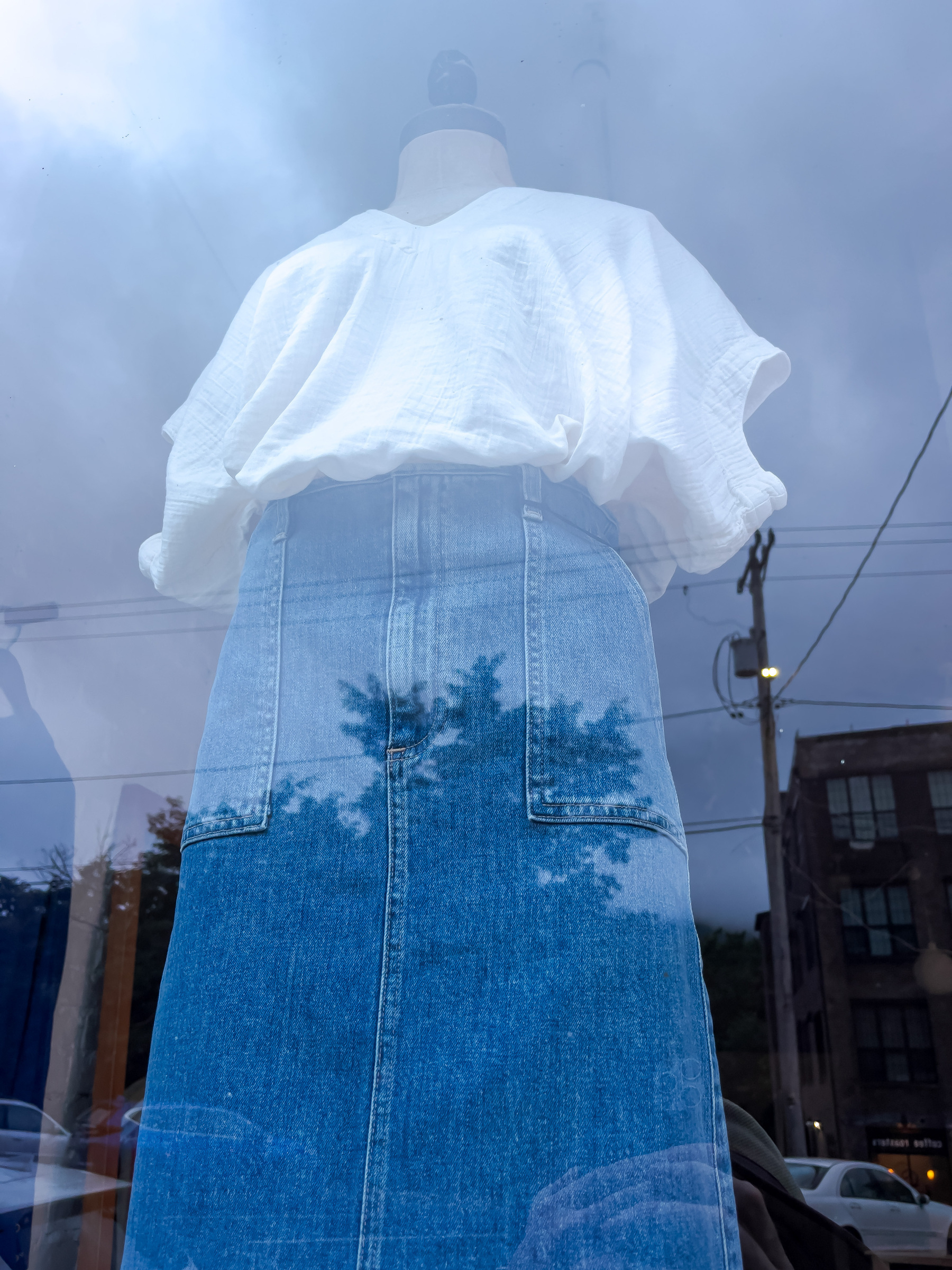 Skirt and simple v-neck cotton blouse on a mannequin in a shop window. Sky and buildings overlay reflected by window.