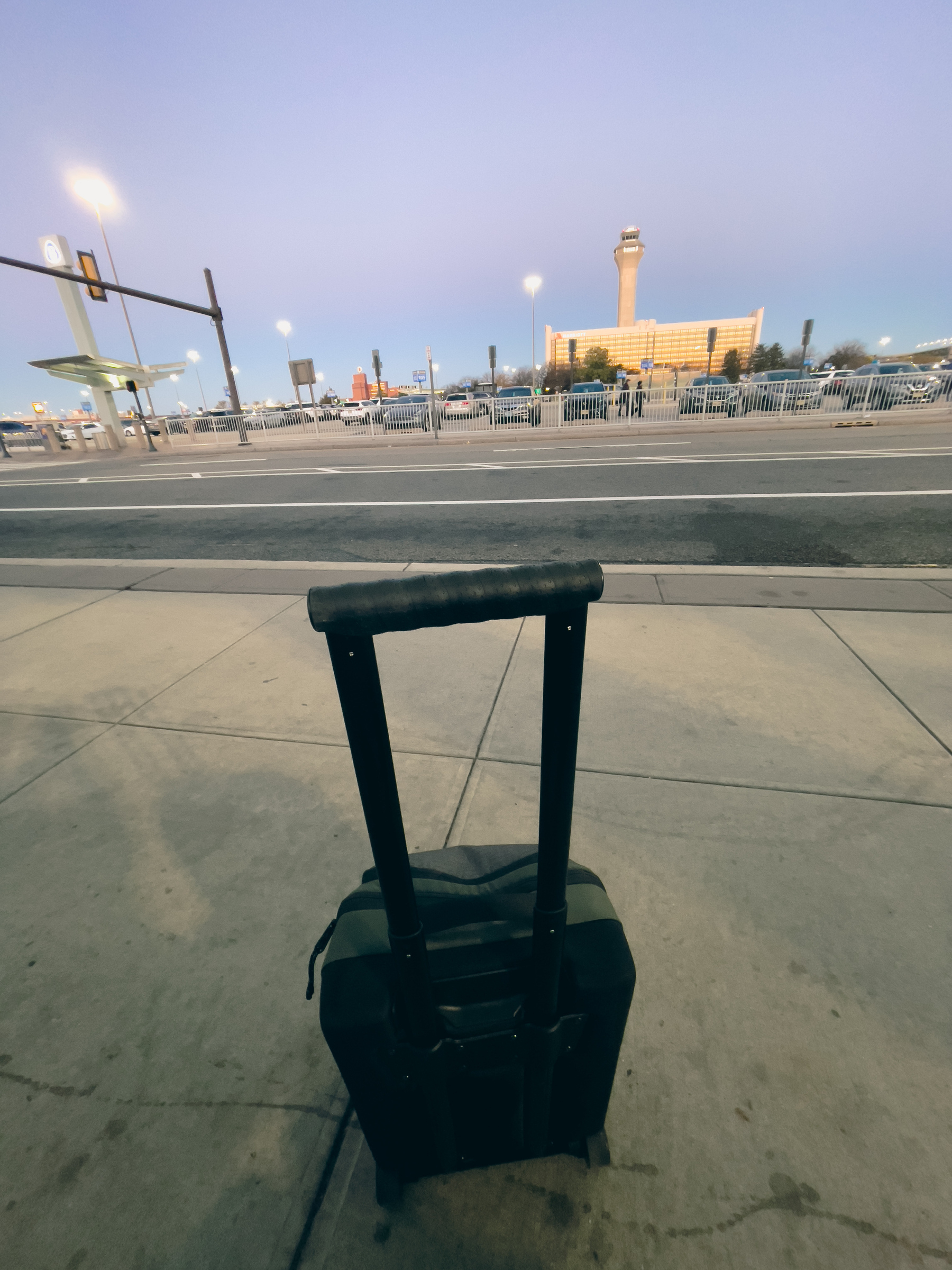 Carry on type suitcase with handle and wheels in foreground, parking lot and control tower building in background.