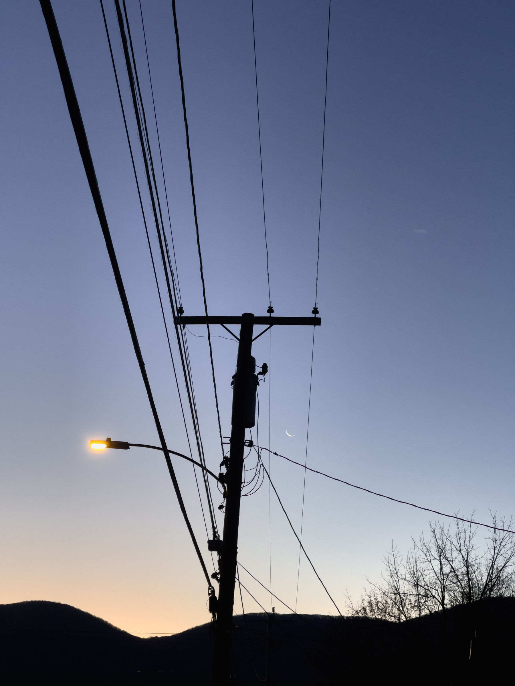 Utility pole in shape of cross with streetlight and wires, sliver moon visible between wires, mountain silhouettes in background at bottom of frame. 