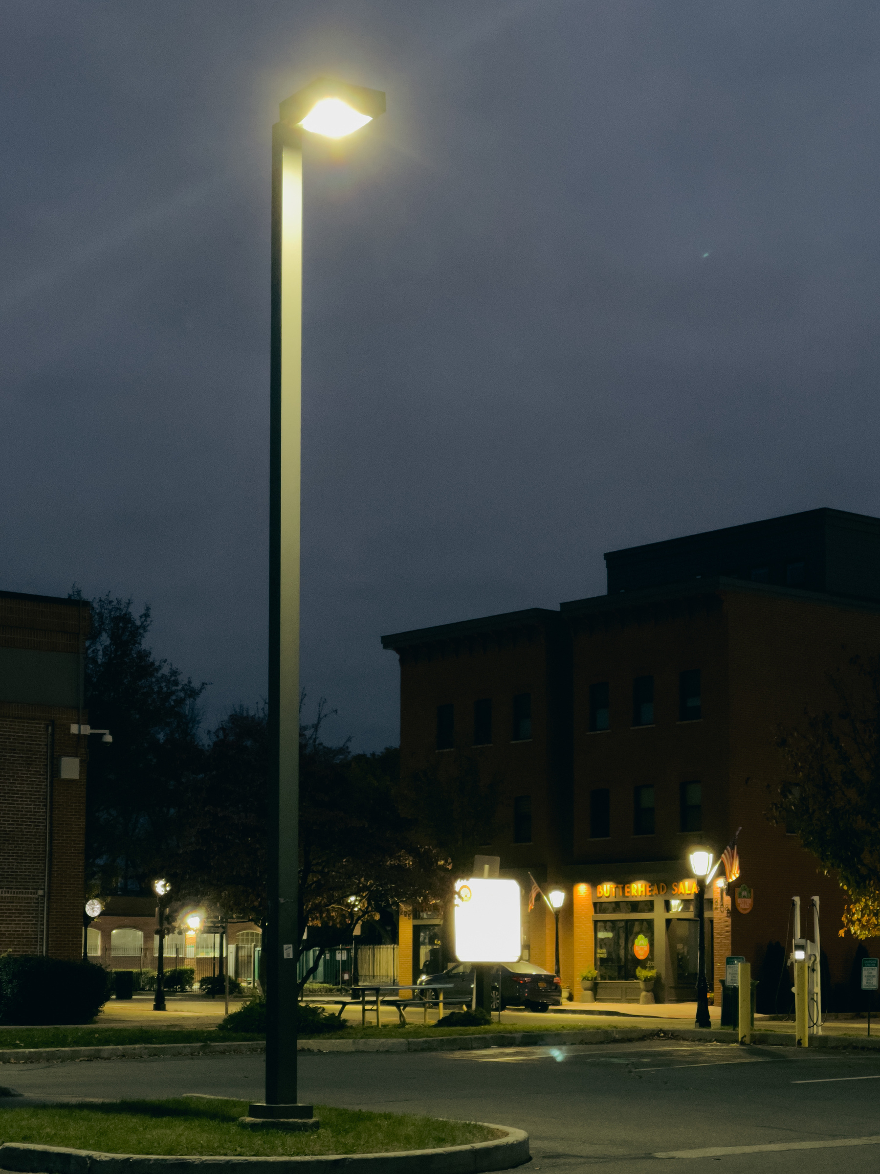 Streetlight in parking lot with silhouetted buildings in background and street lights illuminating ground floor of buildings and street, in background.