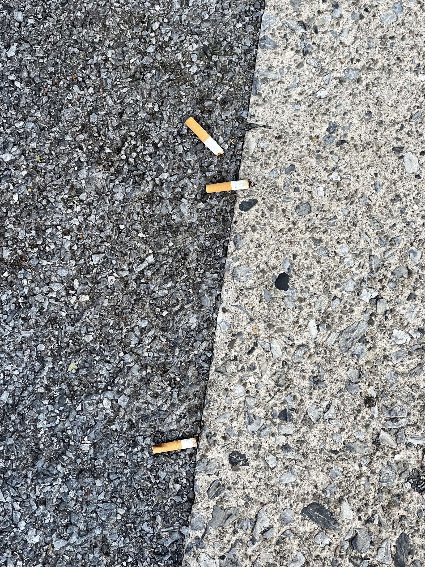 Three cigarette buts on asphalt and concrete pavement. The line between asphalt on the left, and concrete on the right runs at a slight diagonal from top to bottom in the middle. Cigarette buts are arrayed along the line.