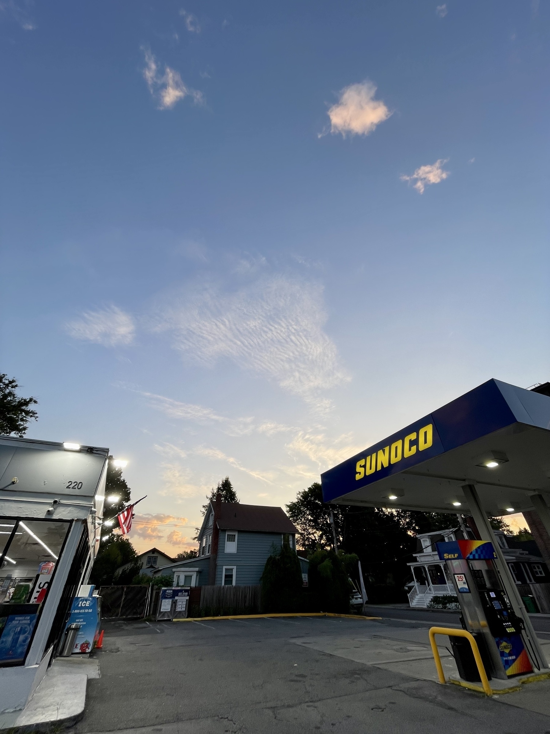 Sunoco gas station pumps on the right, convenience store on the left, sunrise sky above with fair-weather clouds. Made with very wide angle lens.