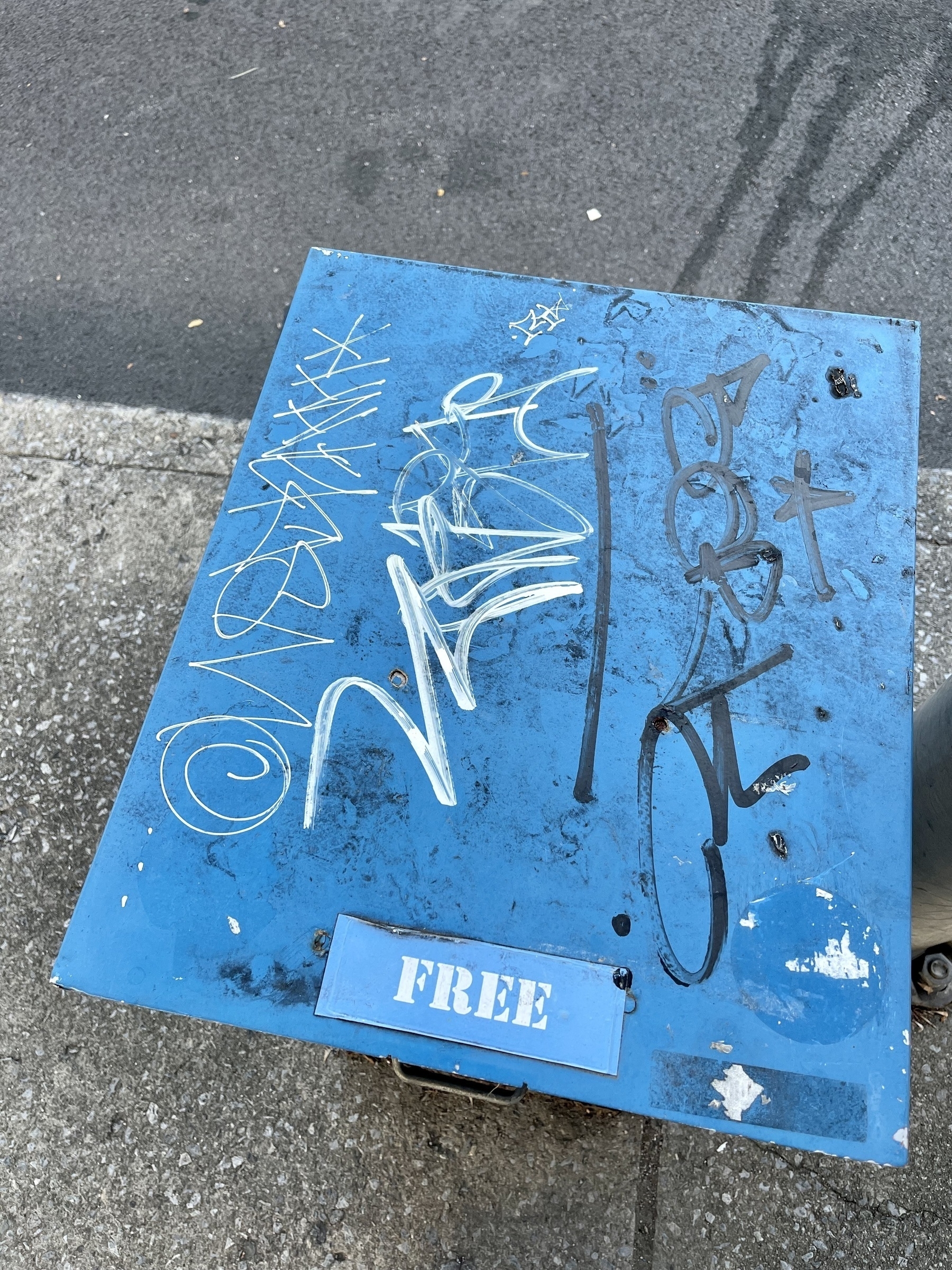 Graffiti on top of blue newspaper dispenser box. A sticker with the word “free” on the lower edge of the dispenser top. Concrete and asphalt paving below.