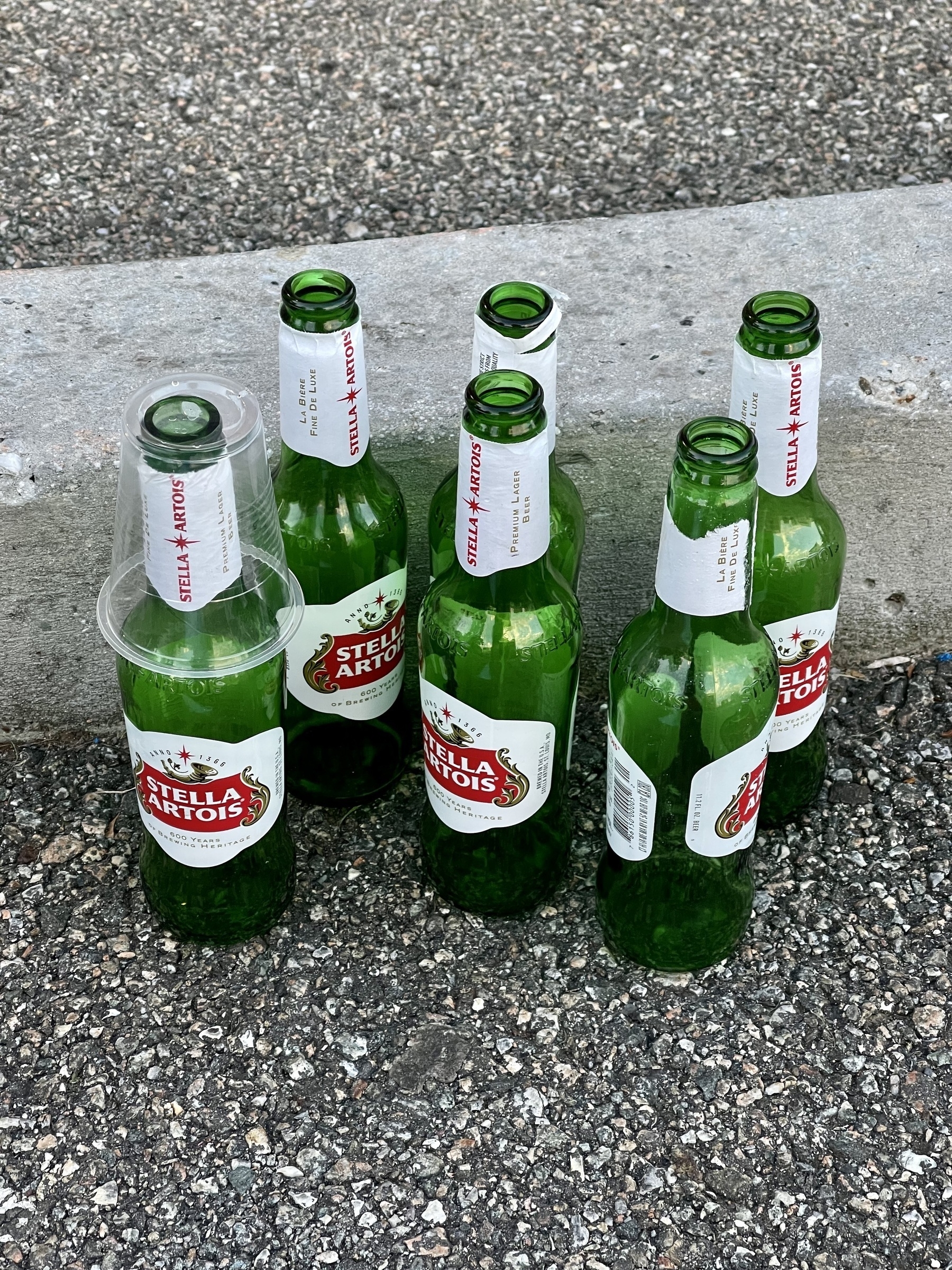 Six empty Stella Artois bottles standing next to a concrete curb in a parking lot. The left most bottle has a plastic cup inverted over it.