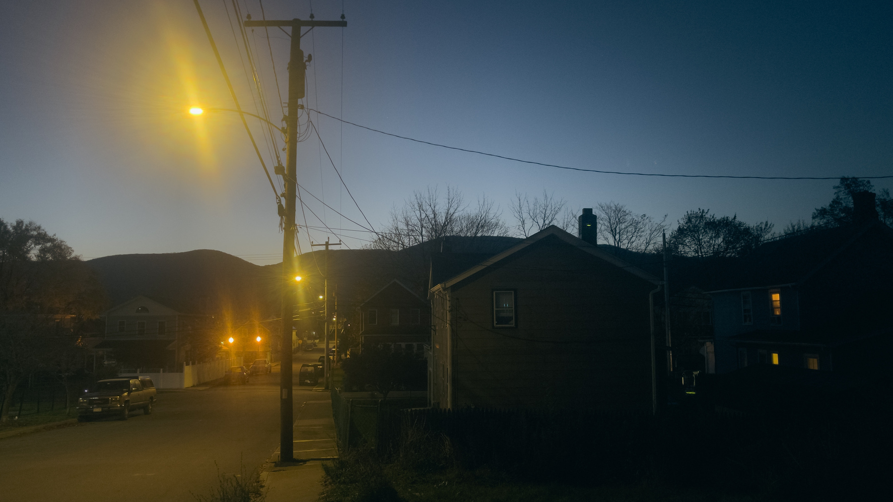 Early morning landscape/streetscape with mountains in the distance, utility poles and streetlights receding into the distance and houses in silhouette to the right.