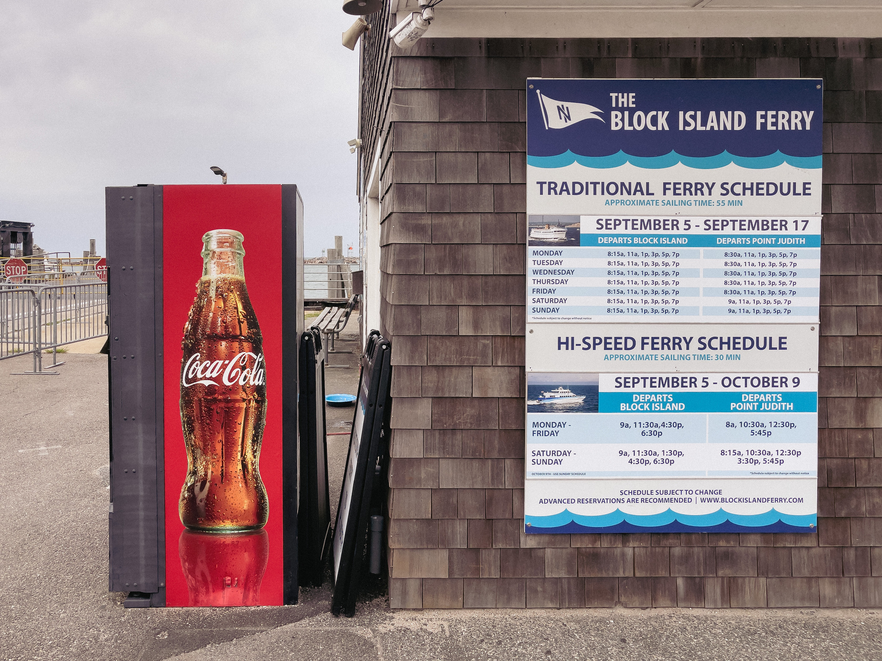 Coke vending machine with large image of a bottle of coke standing next to a building with Block Island Ferry schedule on its side.