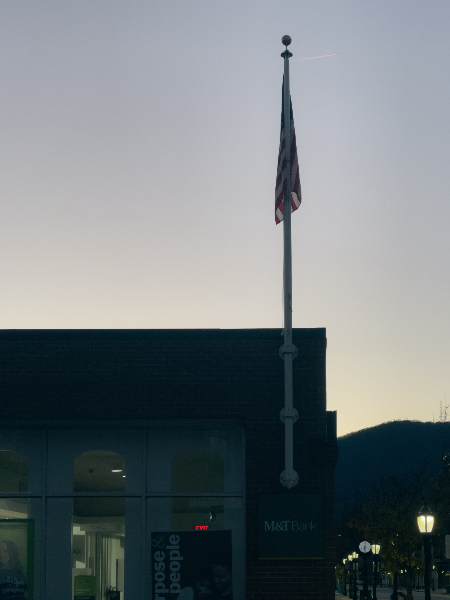 Silhouetted flag pole with American flag mounted on a building also in silhouette and silhouetted mountains in the distance. Dawn sky.