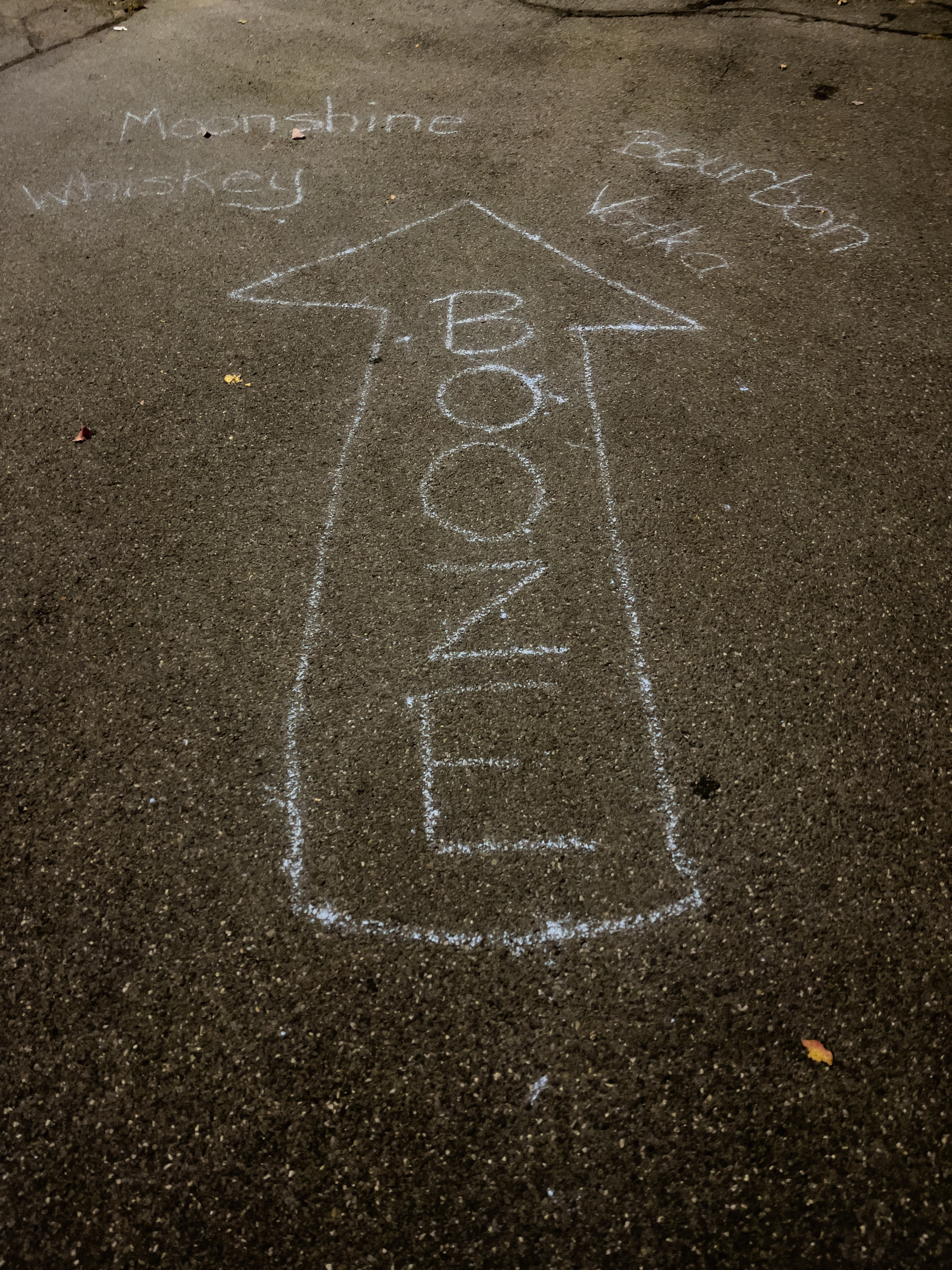 The word Booze inside a directional arrow chalked onto asphalt paving, pointing to the words moonshine, bourbon, whiskey and vodka, also chalked onto the pavement.