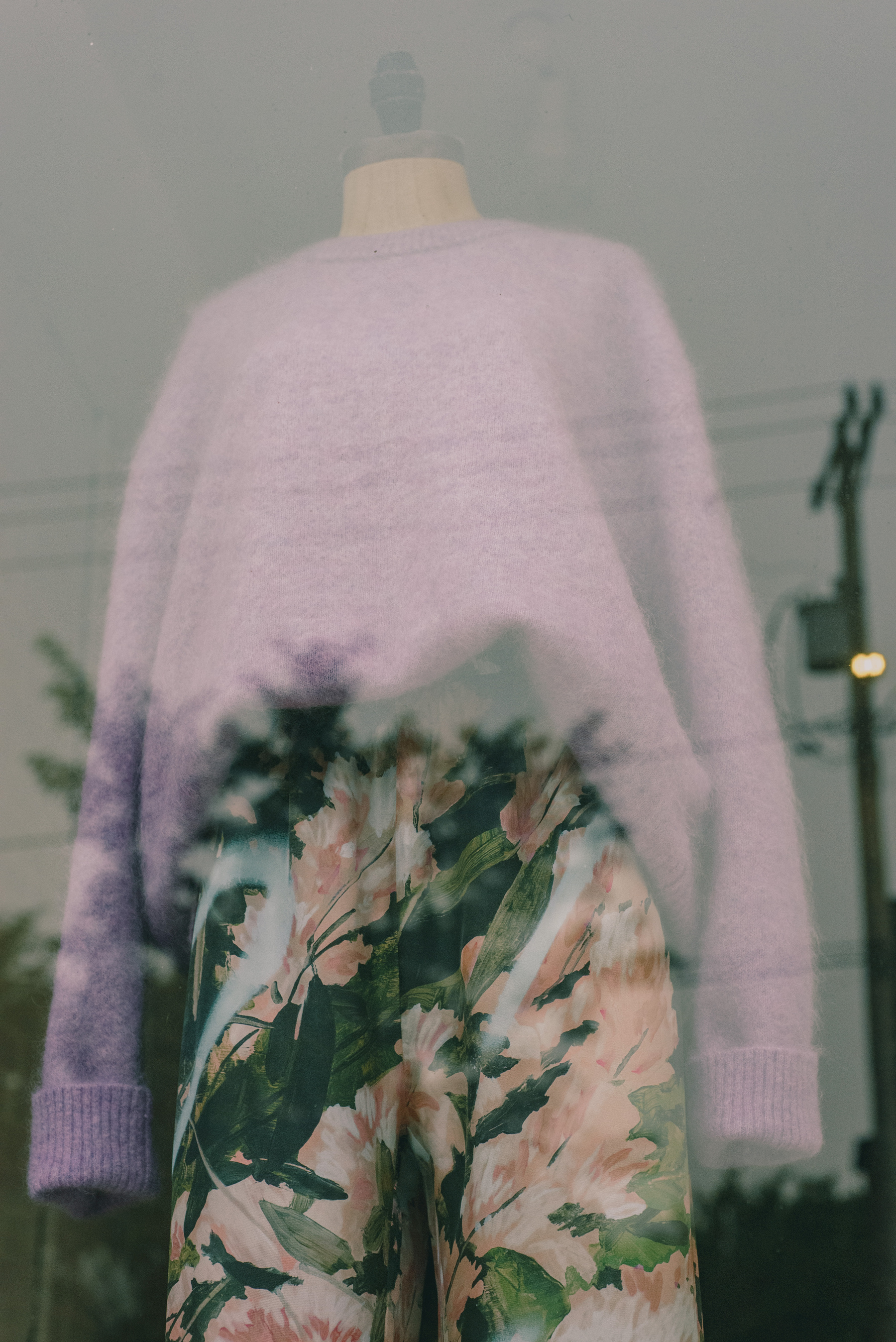 Women’s pink sweater and botanical print fabric pants on a mannequin in a shop building. Wires and trees reflected in window overlayed.