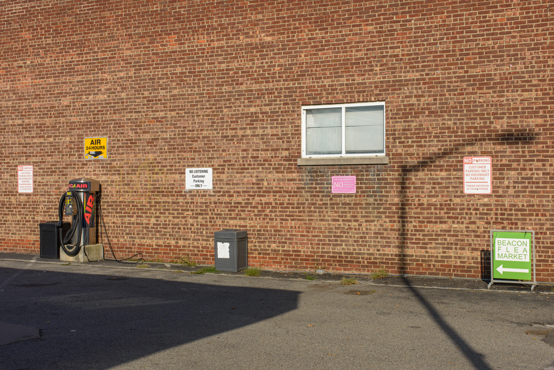 Brick wall with window, signage, car vacuum vending machine, garbage can, on or in front of it. A rectangular shadow of a building projects in from the lower left corner, the shadow of a street light projects in from the lower right corner.