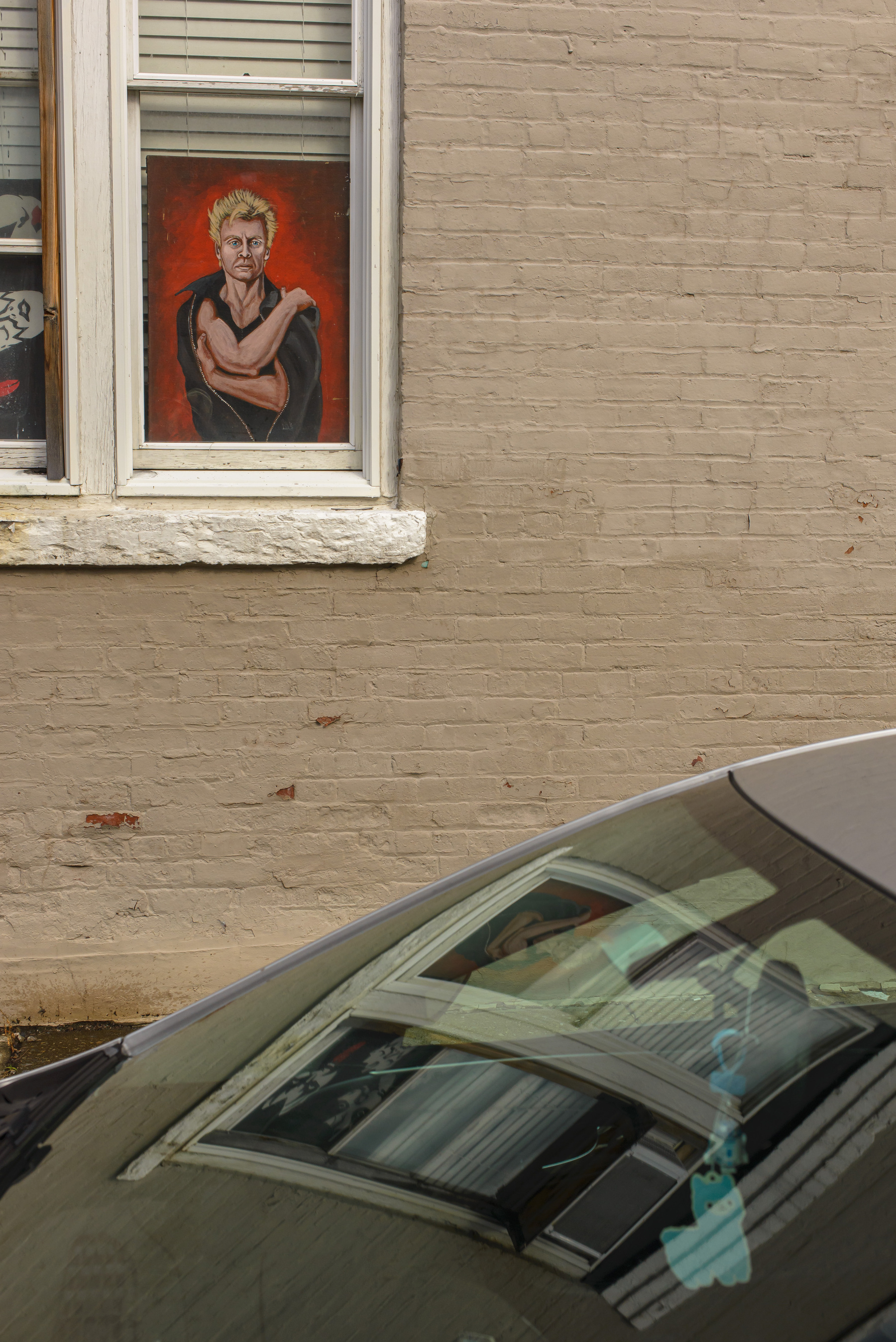 poster of a man in a window in the upper left corner, car windshield across bottom of frame, window reflected in the windshield