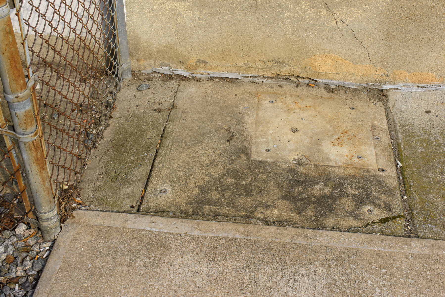 close up of rectangular concrete sidewalk surface with square patch where a pedestal used to be, chain link fence on the lefthand edge of the frame