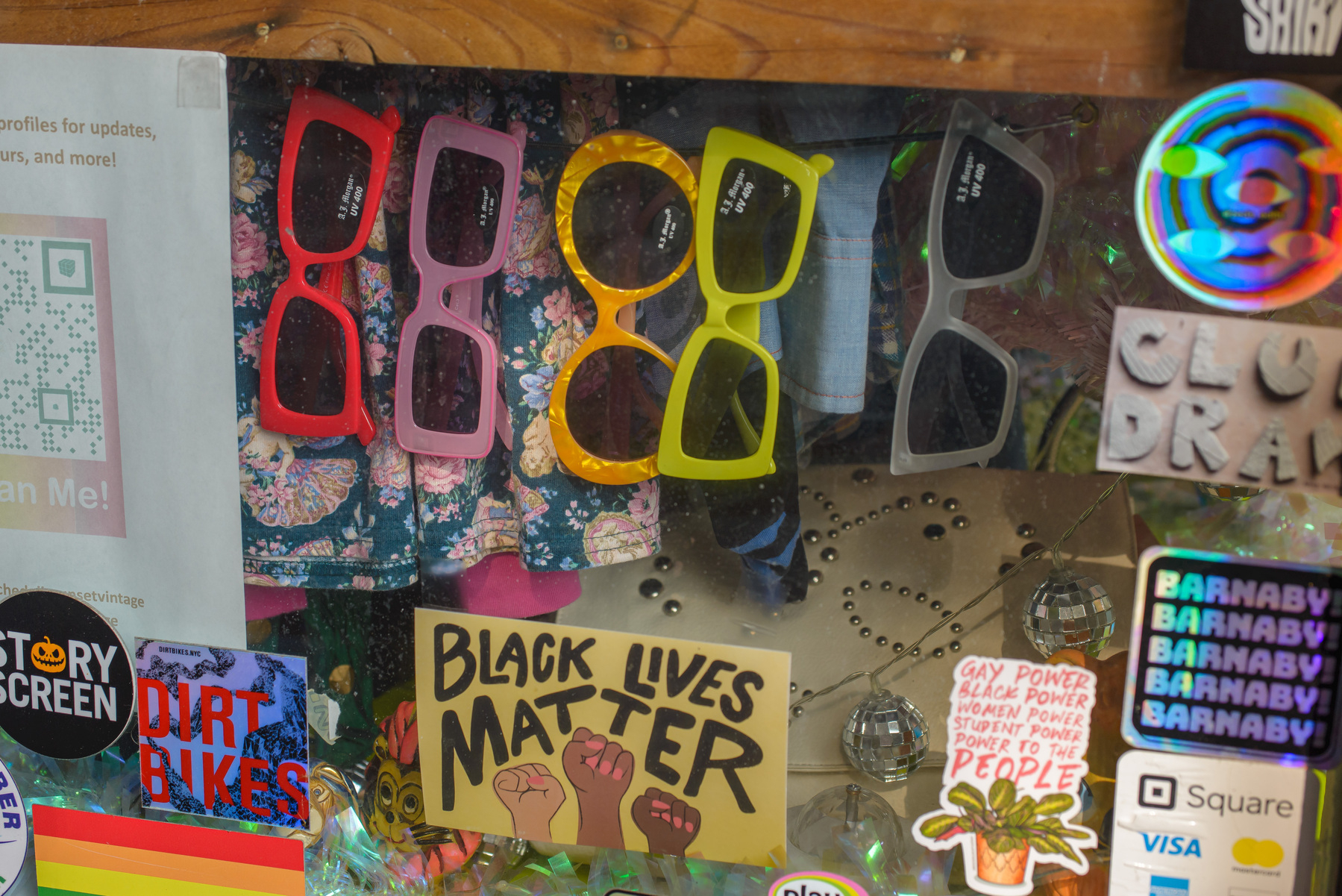 row of colorful bold sunglasses, in a shop window, stickers on the window running around the lower and right edge of the frame, black lives matter sticker with three fists in the middle of the lower edge of the frame