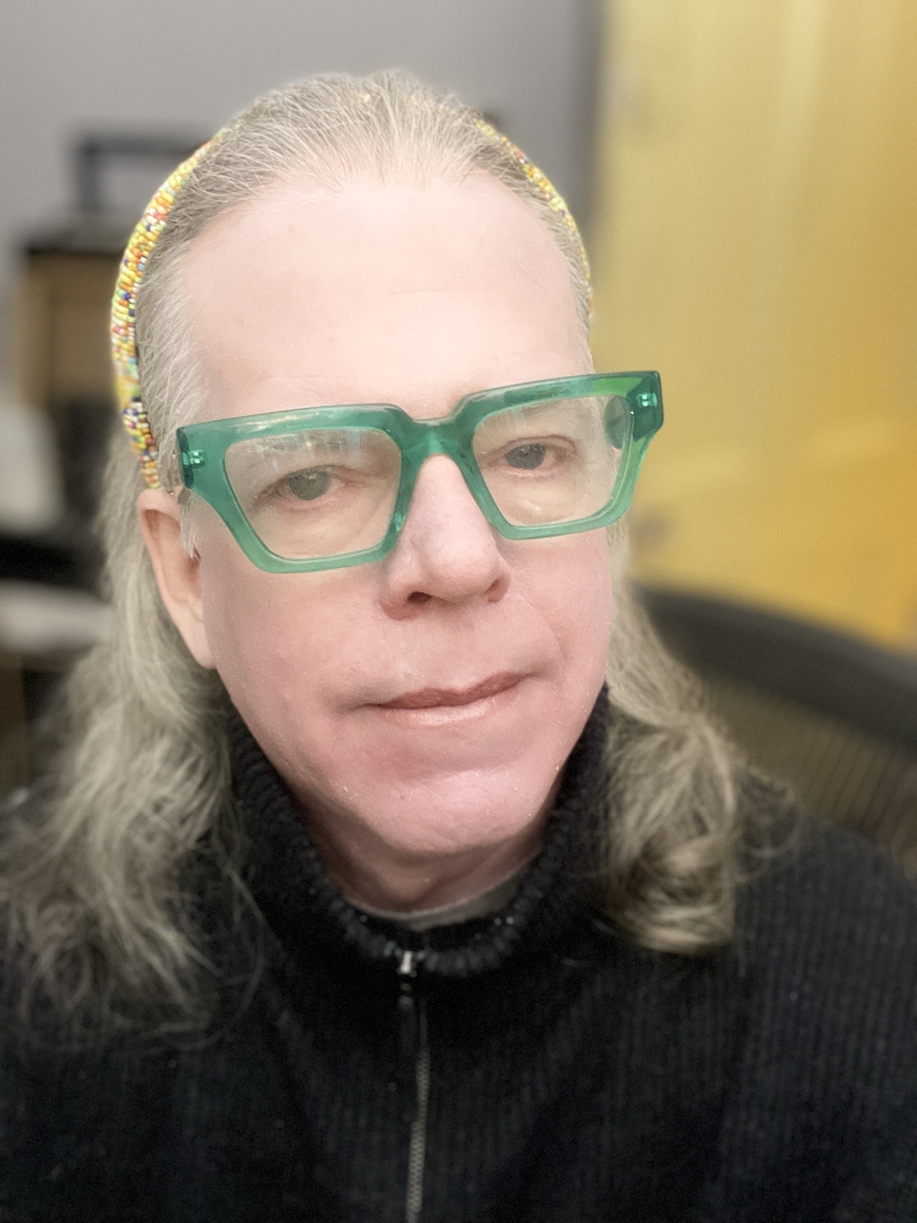 Portrait of trans-feminine man, with multi colored beaded headband, crystal green thick frame glasses and peach colored lipstick.