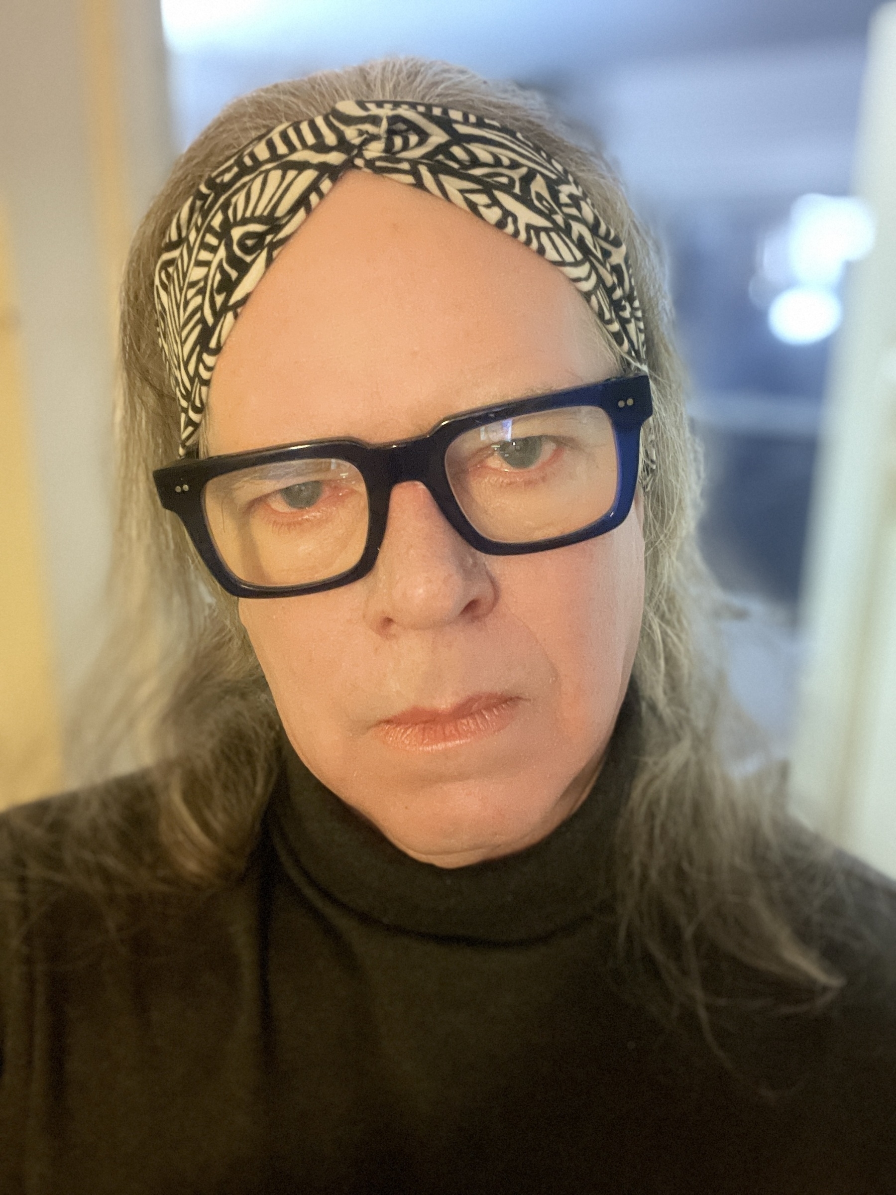 Head shot of trans-feminine person wearing thick framed, dark blue glasses, batik print headband and gray turtleneck shirt. Hair is shoulder length. Her expression is one of slight irritation.