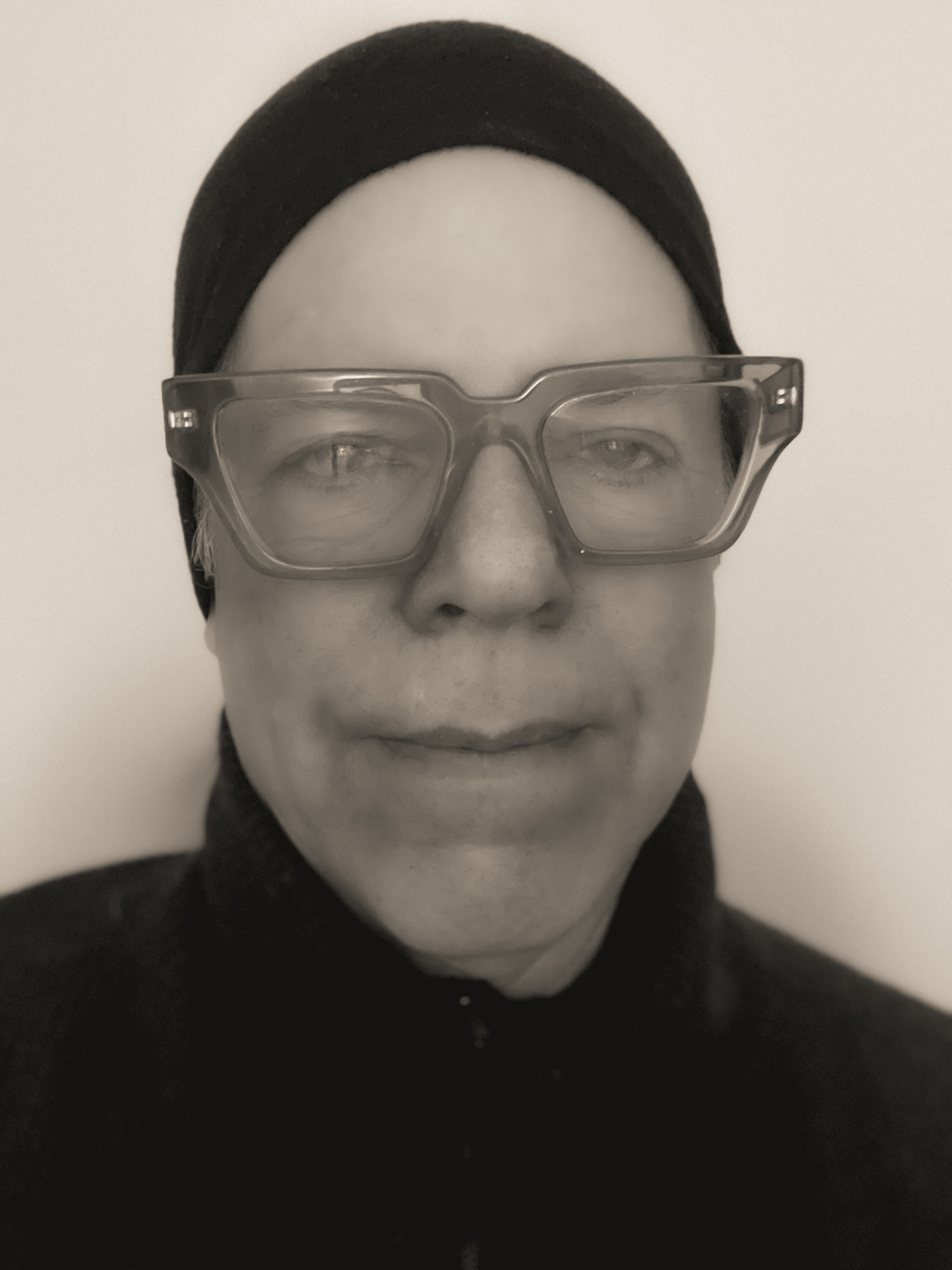 Selfie head shot of artist with prominent crystal frame glasses, black headband and dark grey turtleneck.