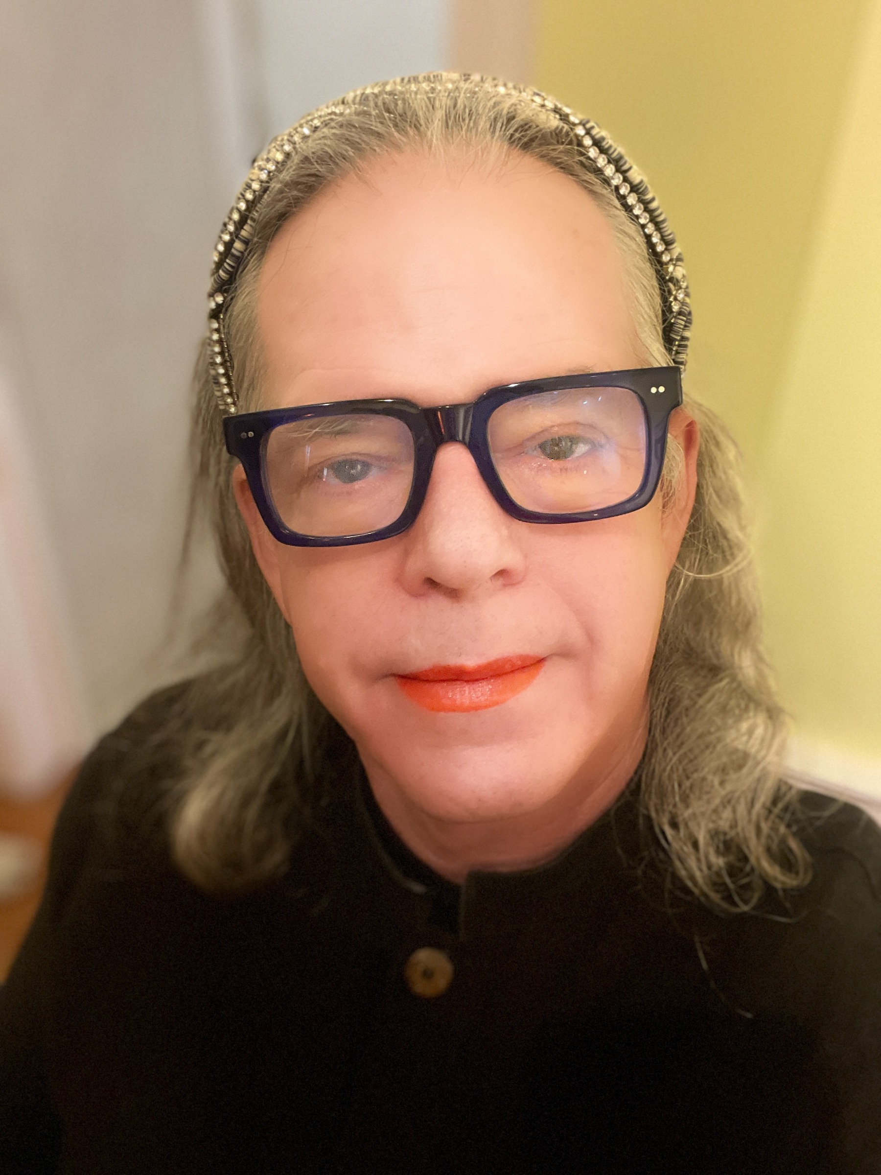 Trans person headshot. Dark blue thick frame glasses, red-orange lipstick, shoulder length hair, diamond studded headband.