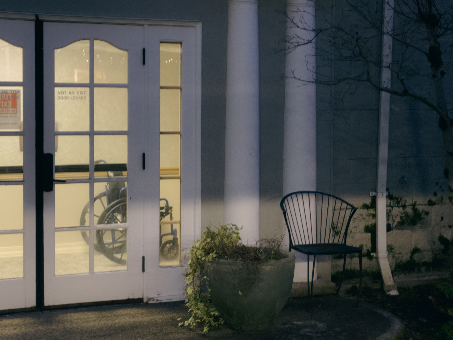 Picture of outside of building. French doors to left. Wheel chair visible through the doors. A garden chair on the outside to the right. A shadow line cutting across scene from left to right.