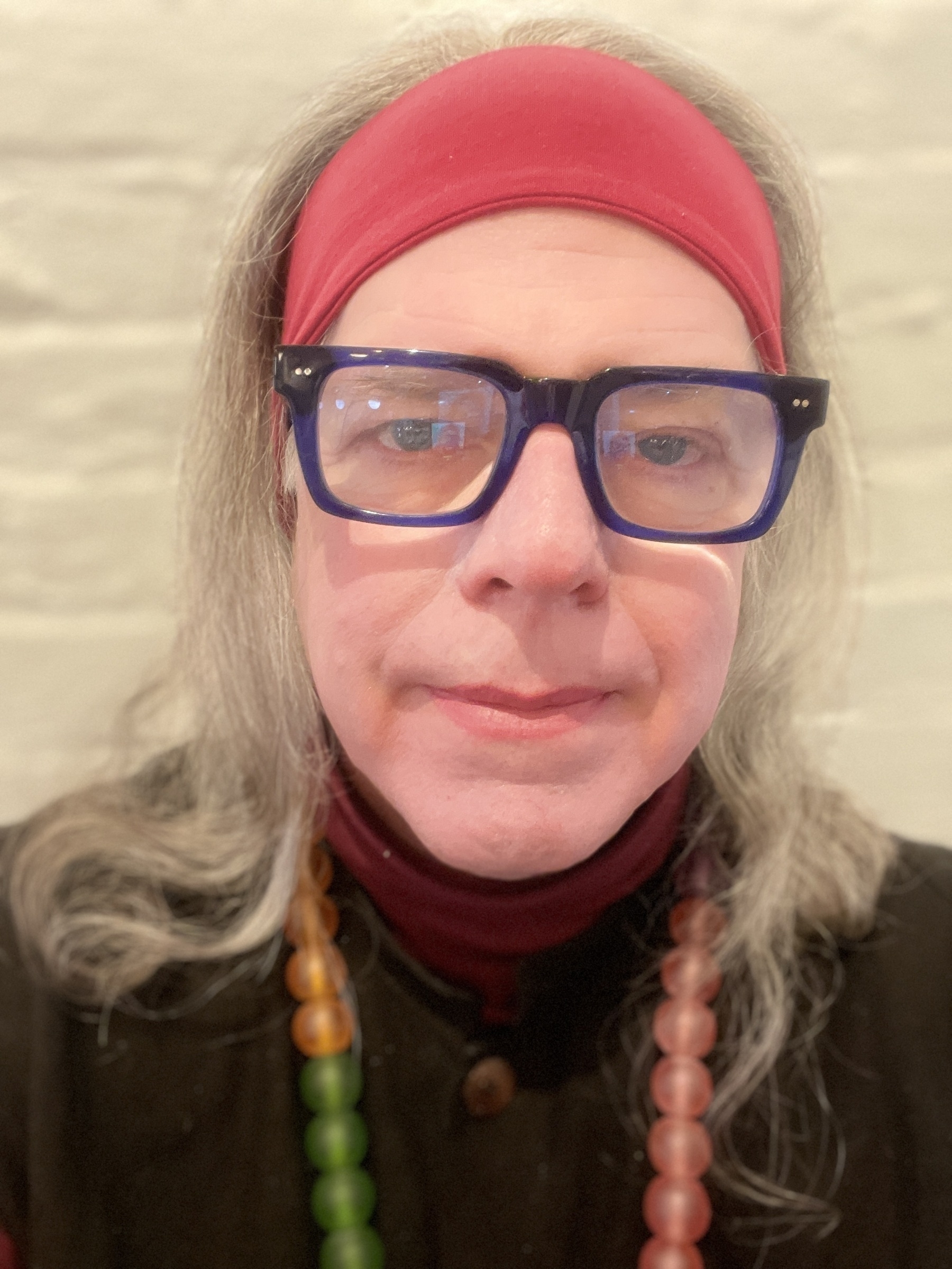 Self portrait trans-feminine man with hair down to shoulders, colored translucent bead necklace pink headband, blue frame glasses and pink lipstick.