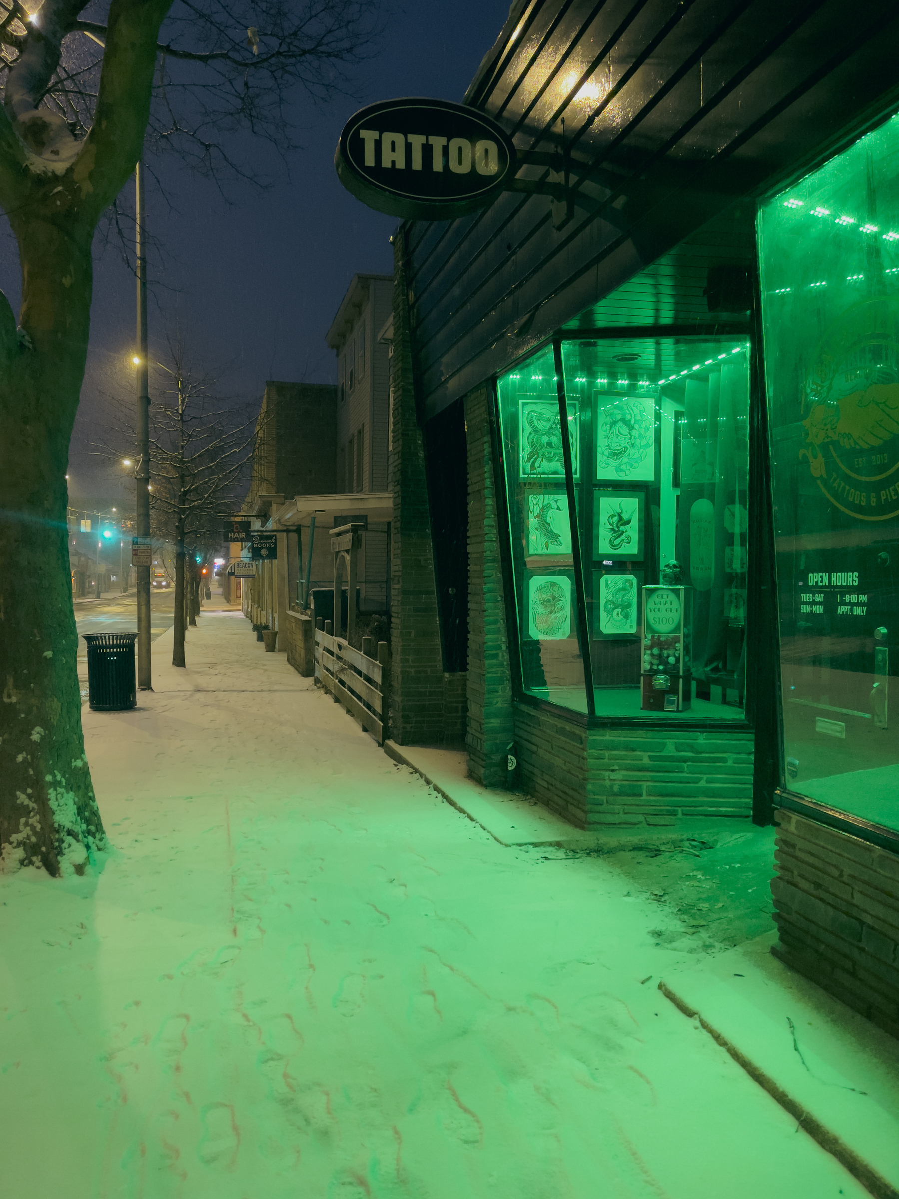 Streetscape looking down snow dusted sidewalk in front of a tattoo parlor storefront with green light from the parlor illuminating the scene.