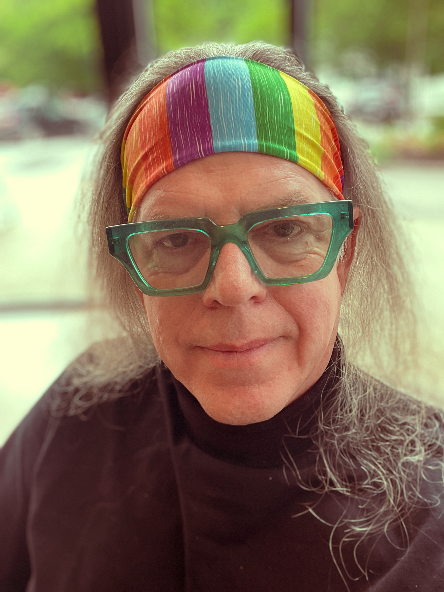 Headshot of a man wearing a rainbow striped head band. He has hair down to his shoulders and bold green crystal frame glasses on. He is wearing a black turtleneck.