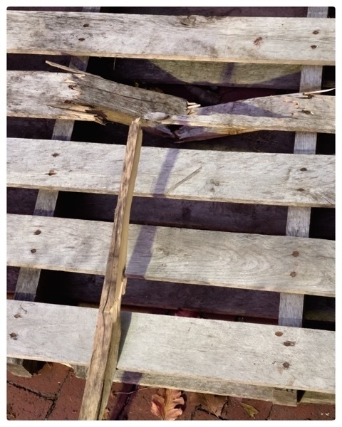 Wood shipping pallet closeup. Paletboard is broken at the top. Woodpiece lies against the pallet and points to the broken board.