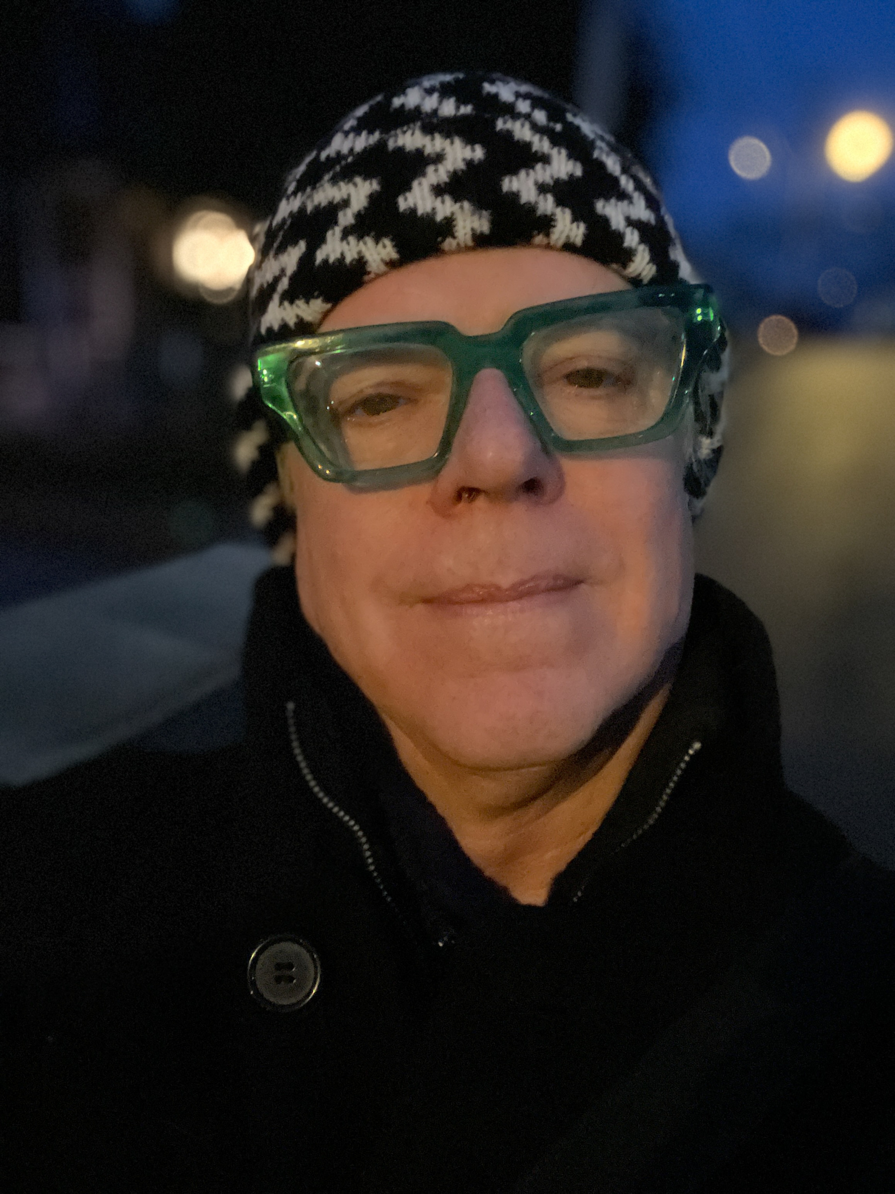 Self portrait with black and white zig-zag knit cap, green crystal frame glasses, illuminated by streetlight.