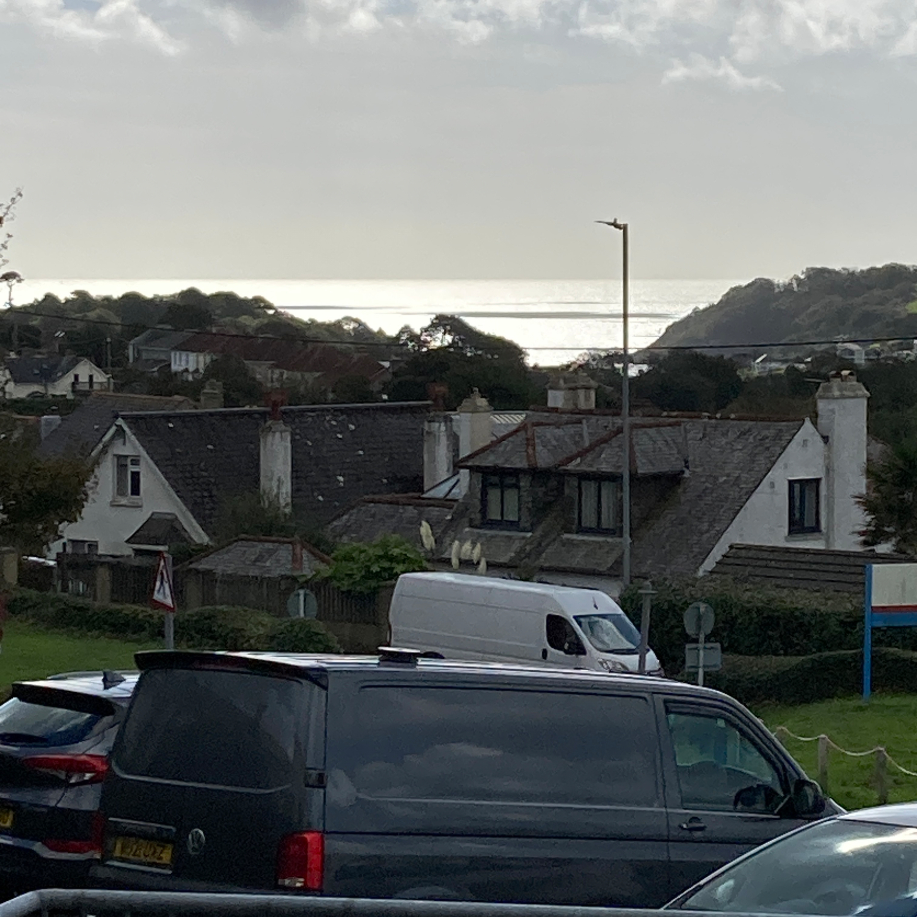 A coastal village scene with houses, parked vehicles, and a body of water reflecting sunlight in the background under a partly cloudy sky.