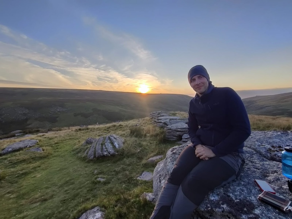 A man wearing a beanie sitting on a rock on a moor with the sun setting behind a hill behind him
