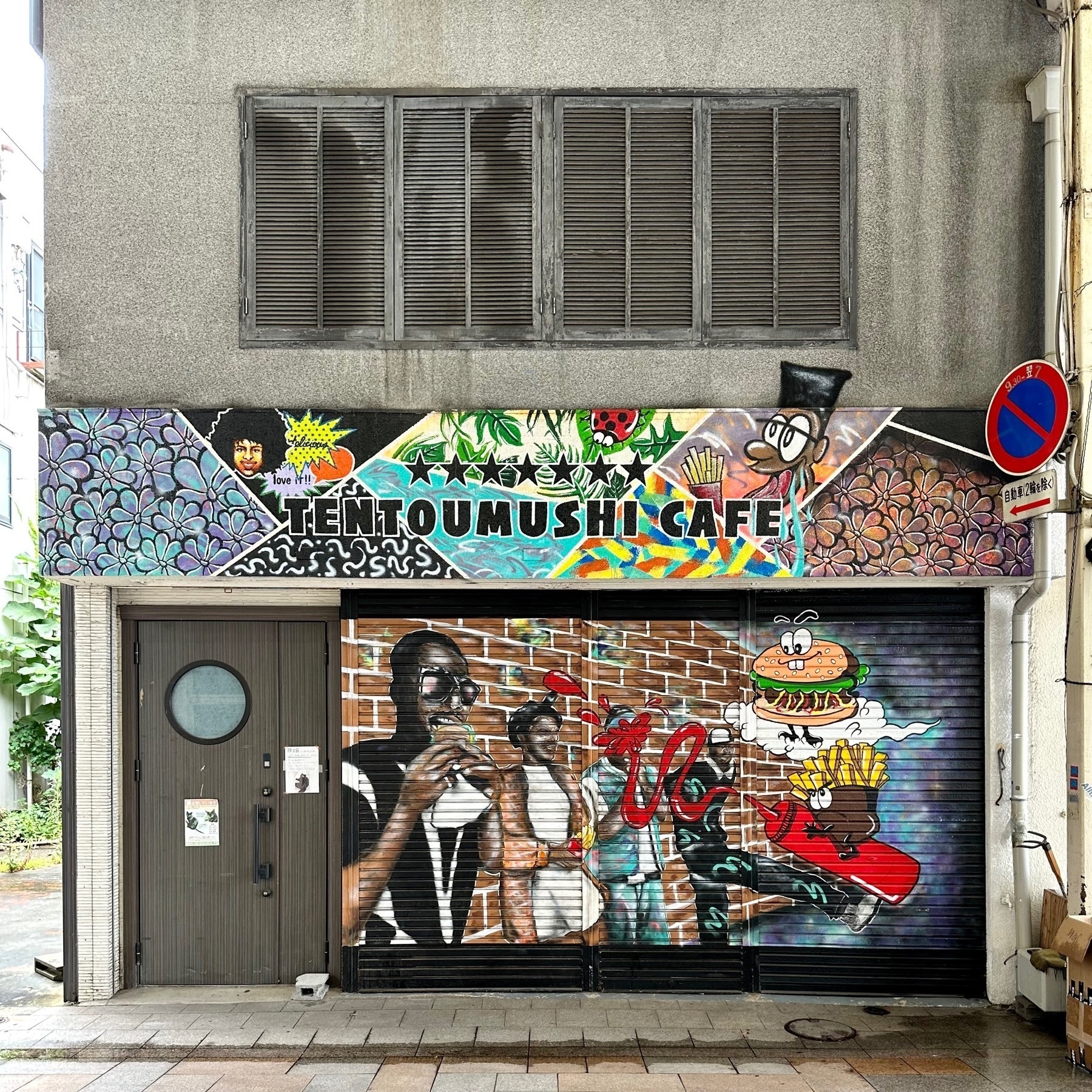 Closed storefront of a cafe called TENTOUMUSHI, meaning ladybird/ladybug in Japanese, with a mural on the shutters of several people eating, a burger, and fries spraying ketchup