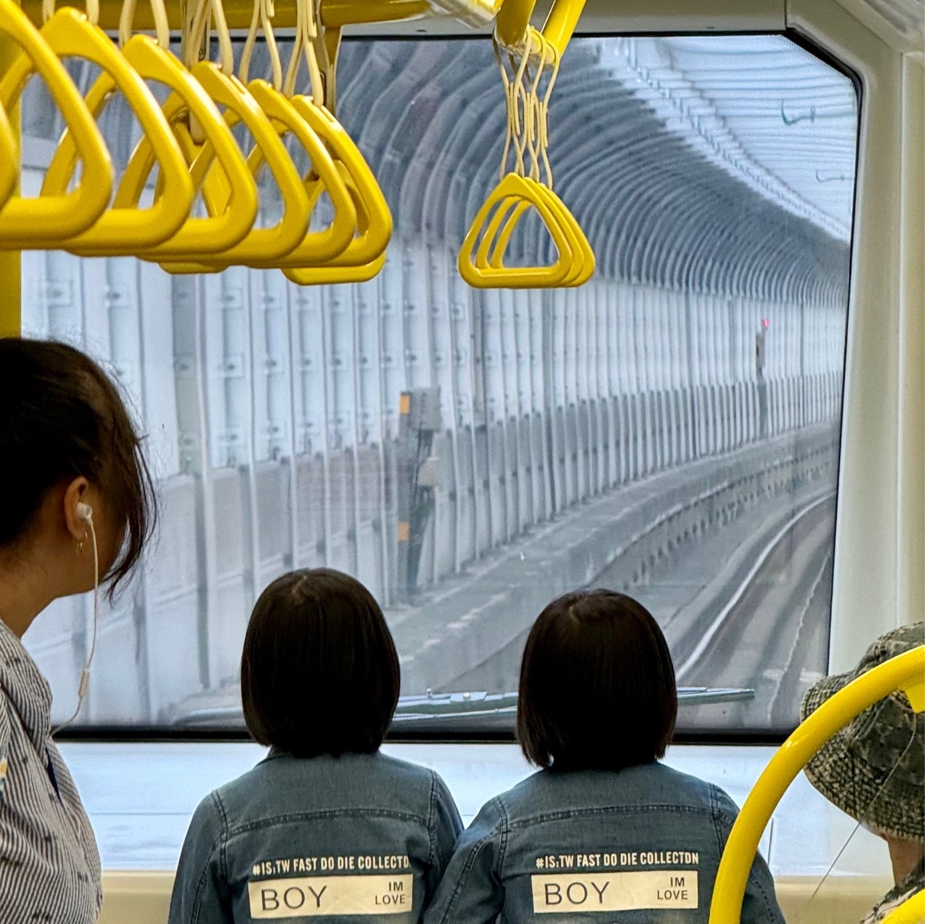 The heads of twin girls with bob haircuts looking out of the front of the (automated) metro train down the tracks, other passengers either side, and many yellow grab handles overhead