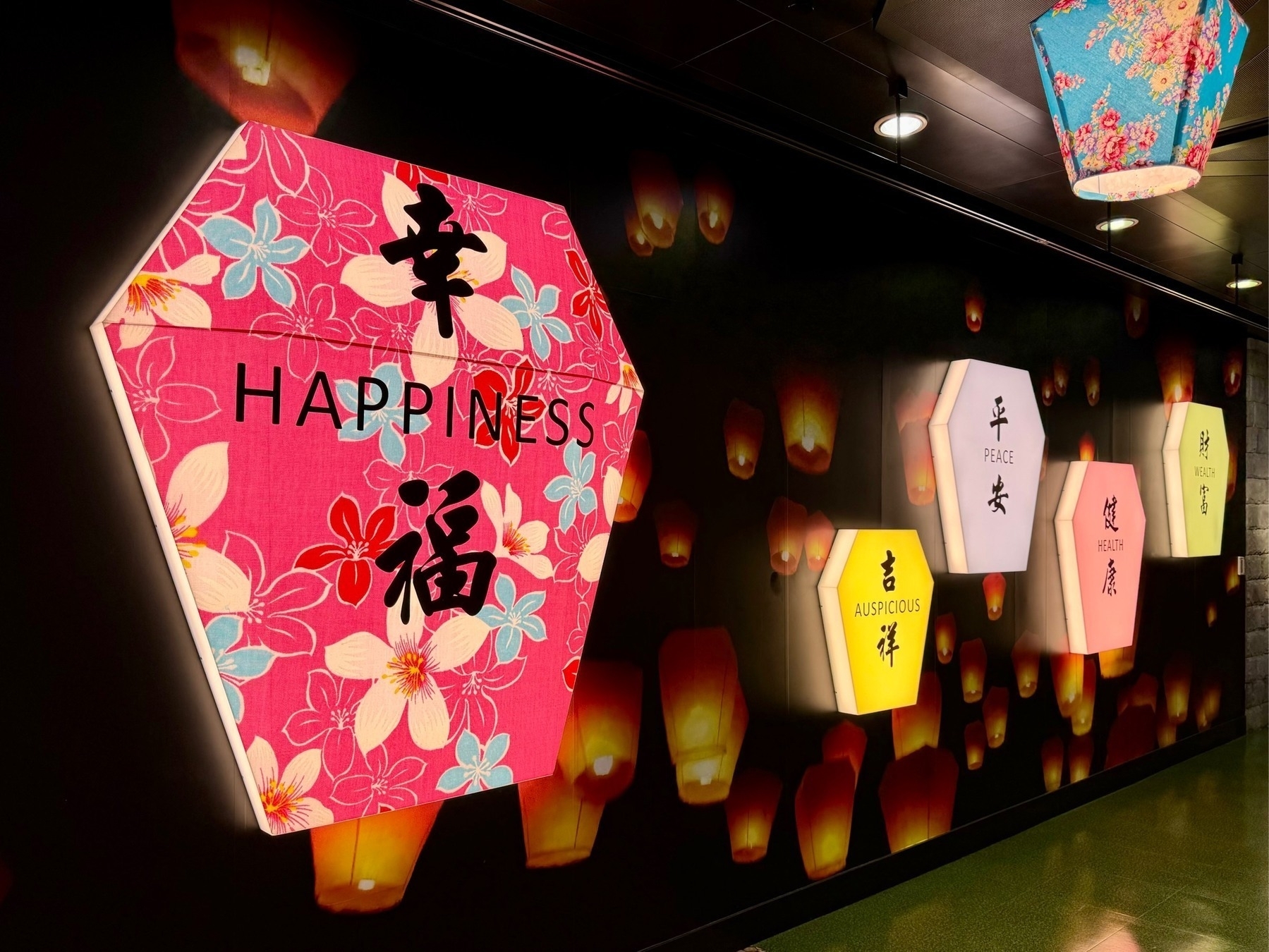 A lighted wall design of sky lanterns, the largest and closest in pink with flowers, having the English word Happiness and the associated Chinese characters