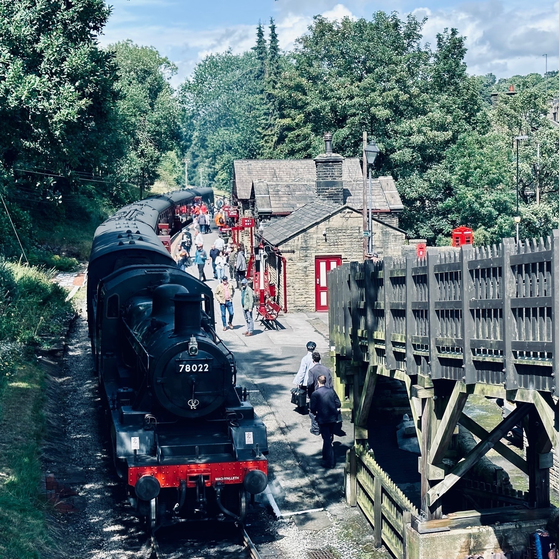 The front of a steam train pulled into Haworth station, people on the platform. Taken from an elevated bridge