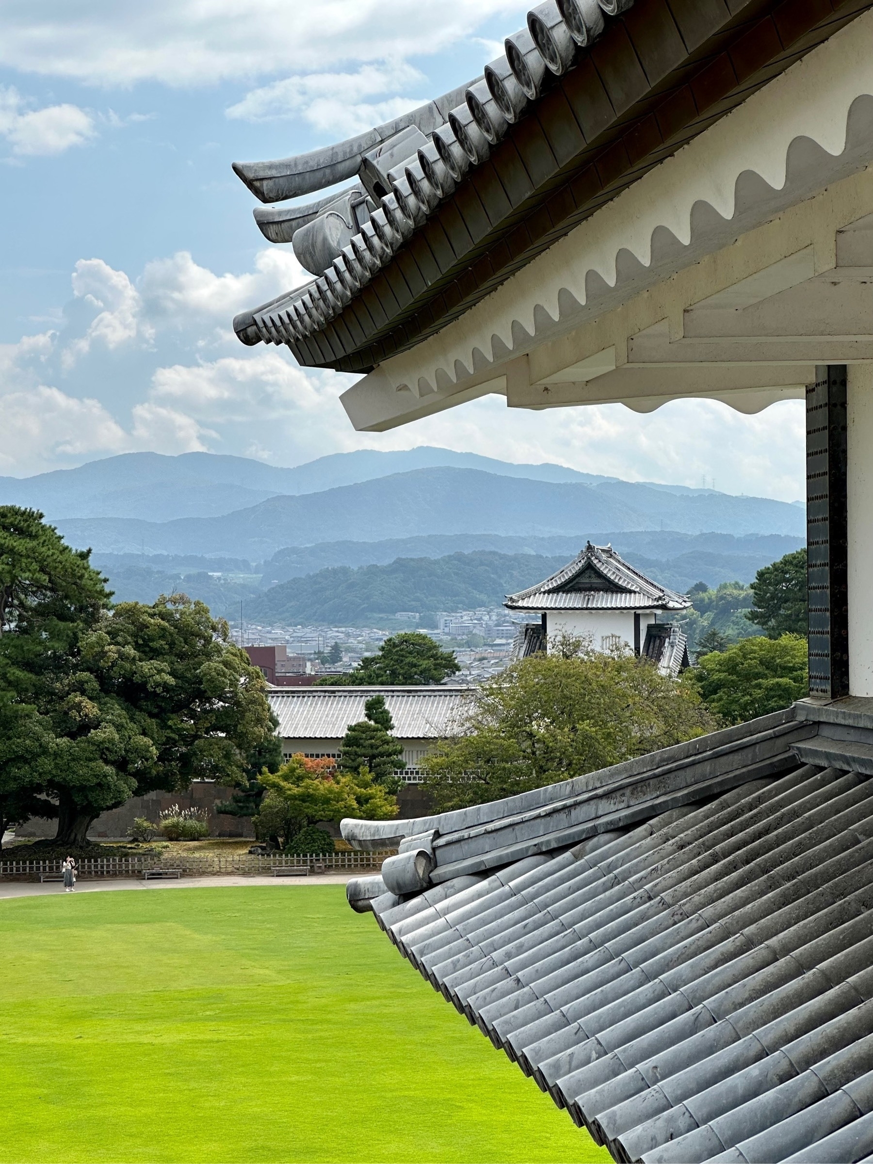 Several layers of misty mountains in the distance, taken from between two roofs of a Japanese castle, a cloudy sky above, and an almost luminous-green field of grass below