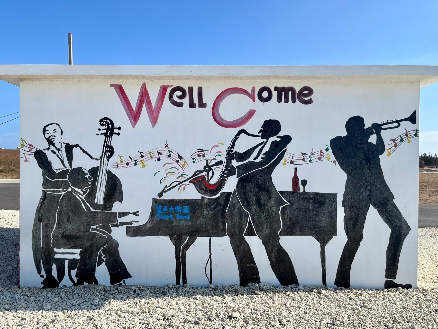 Mural on the side of a low beach-side building depicting a jazz band called "Shark Band", with double-bass, piano, sax, and trumpet, all painted in black, with the words "Well Come" above it