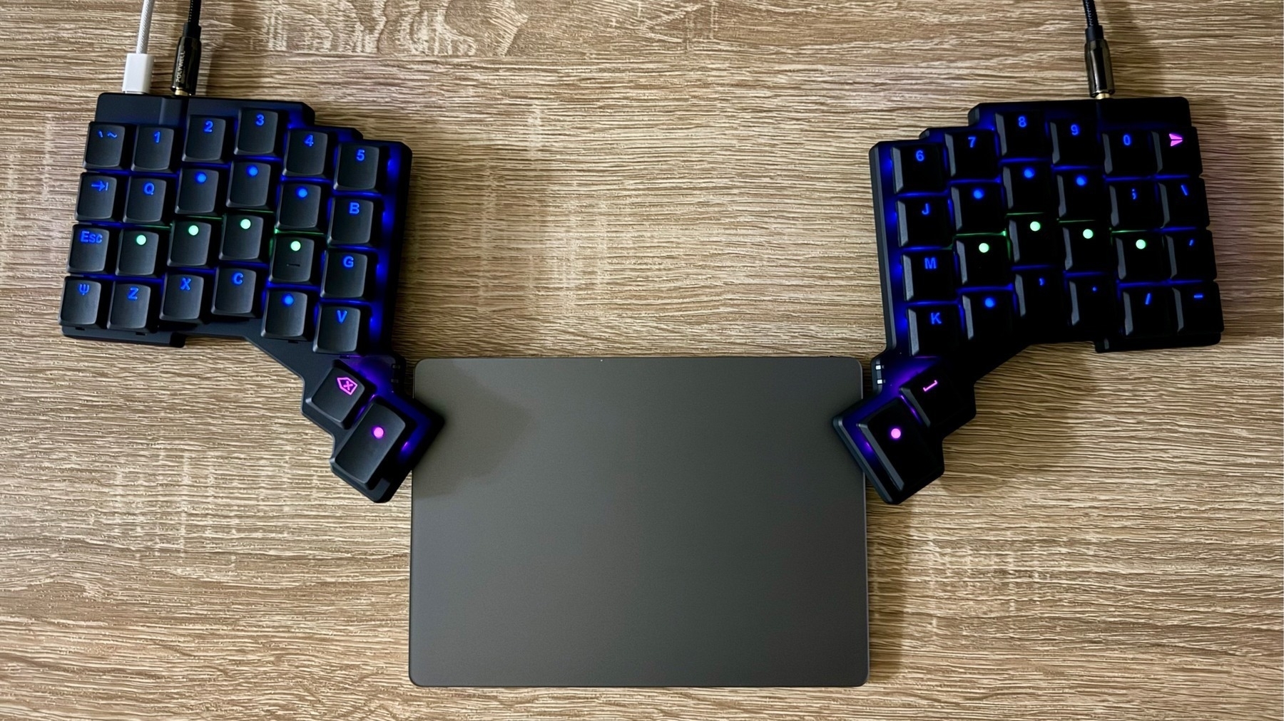 A top-down desk shot showing a black split ergonomic keyboard with RGB lighting, the two halves placed either side of an Apple Magic Trackpad