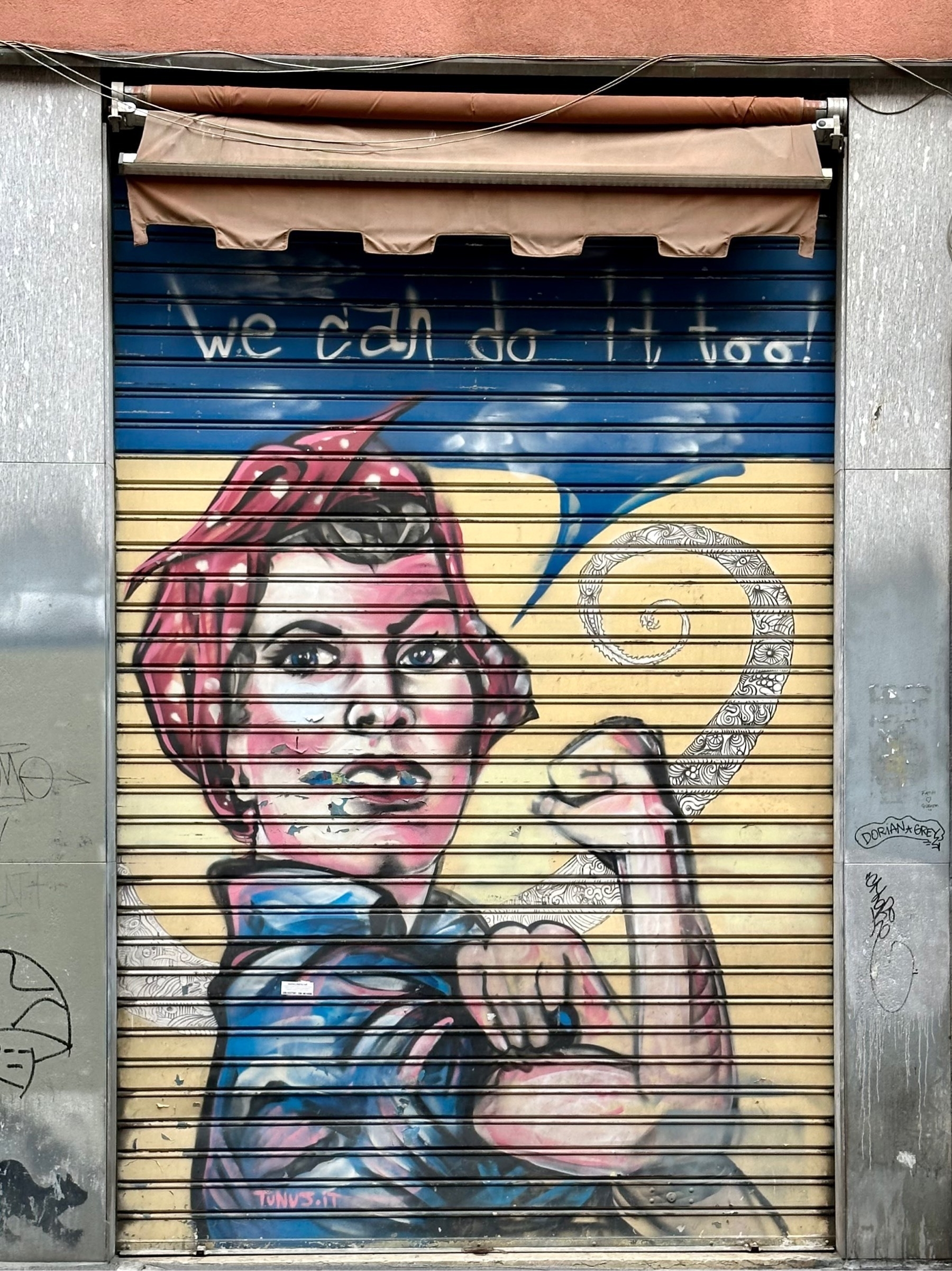 Mural of the text "We can do it too!" above a female caricature in a headscarf flexing her bicep