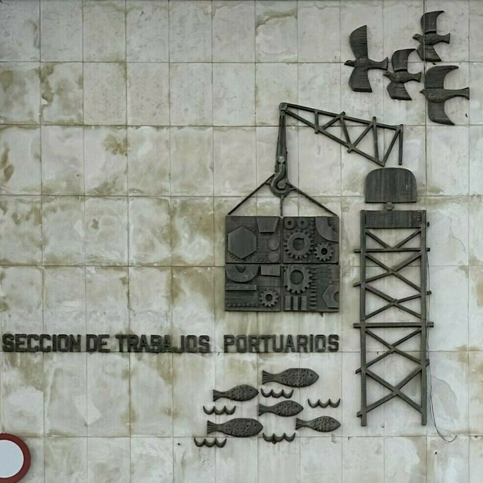 Design on a wall made from pieces of metal, showing a crane with birds and fish, and with the text (in Spanish) Sección de Trabajos Portuarios