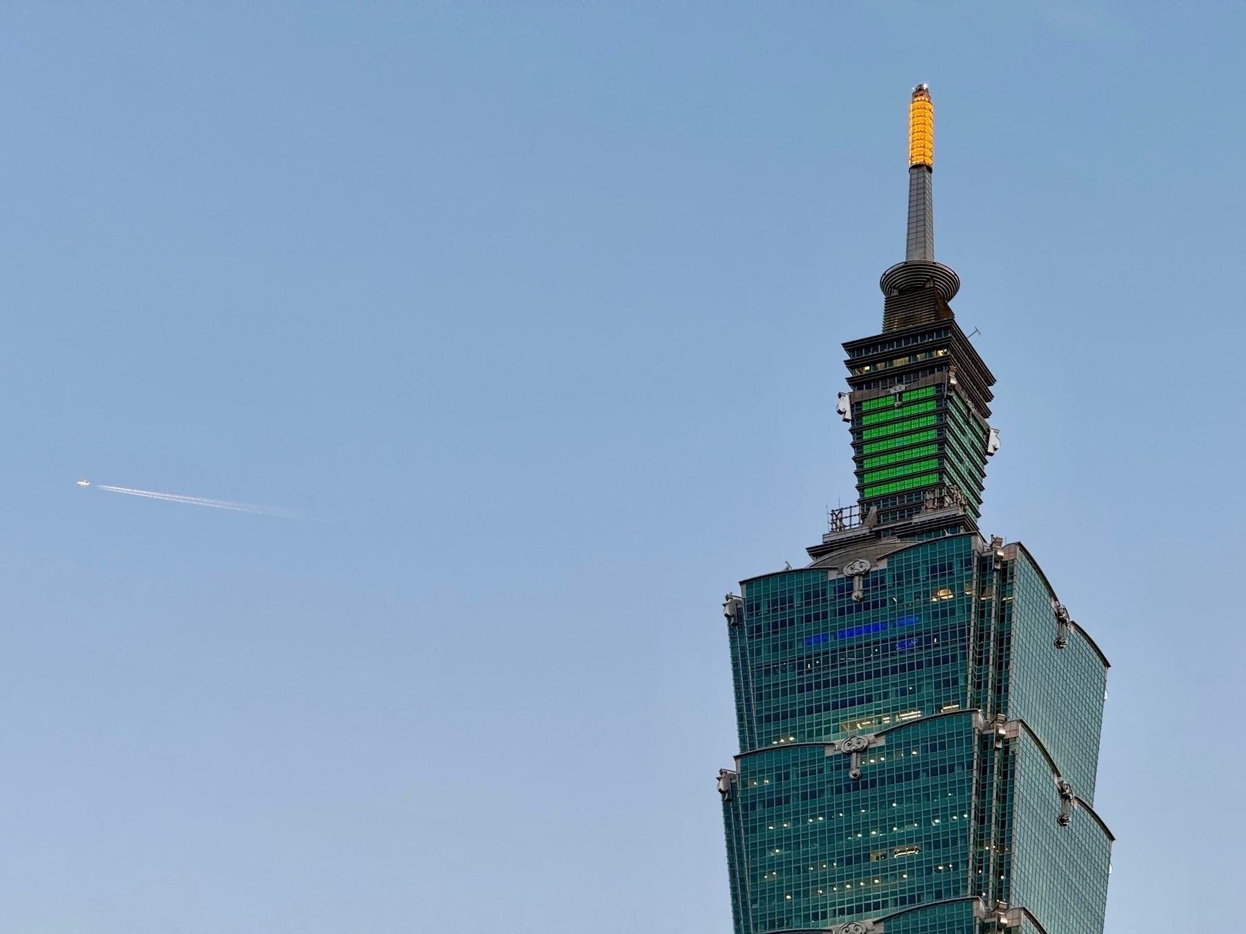 A bright aeroplane being hit by the sun, with a short contrail behind it, having flown behind the top part of Taipei 101, partly lit up with lights around dusk